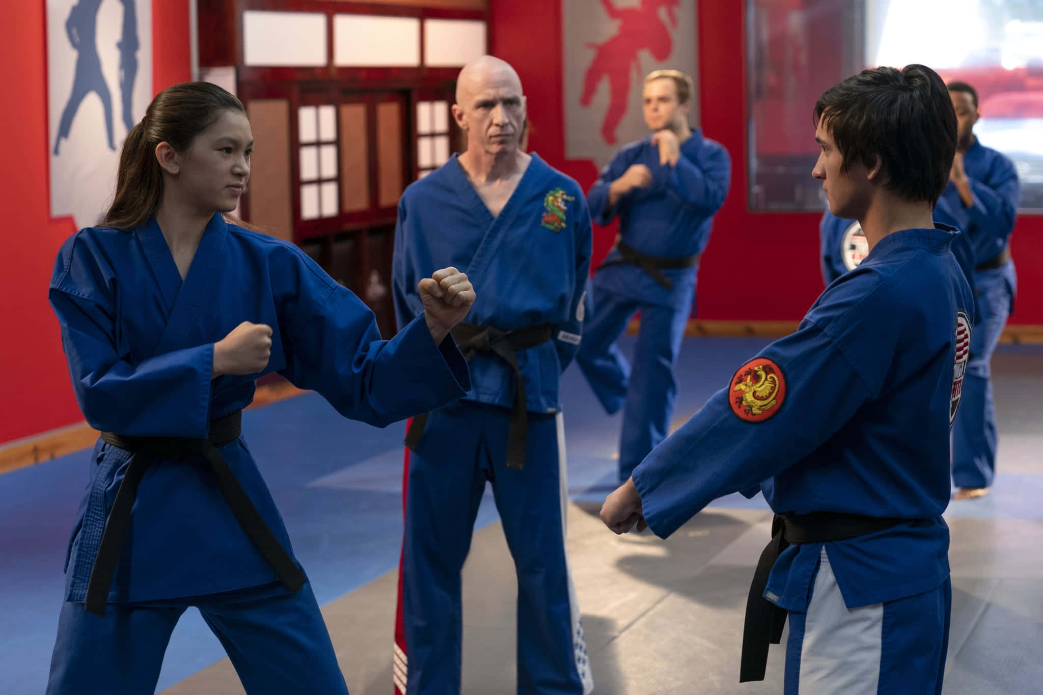 加入開創一切的道場：Cobra Kai！ 壁紙 — 兩名身穿藍色道服的武術家在道場中對峙，一名光頭教練在訓練期間專注地觀看。