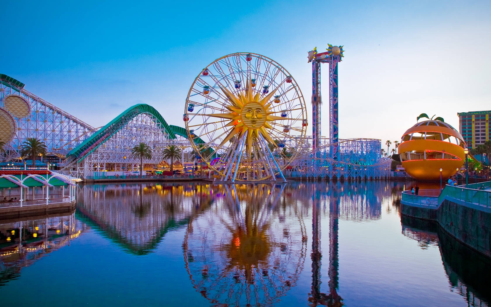 加州冒險 Anaheim 壁紙 — 黃昏時分充滿活力的遊樂園，金色的太陽形摩天輪和過山車倒映在平靜的水面上。