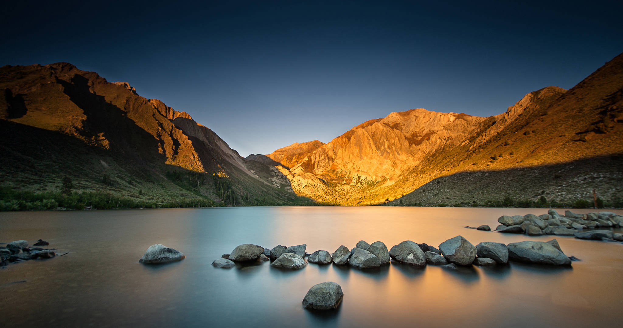 加州因約國家森林 壁紙 — 金色夕陽沐浴崎嶇山頂，溫暖光線反射在平靜湖面，前景散落岩石。