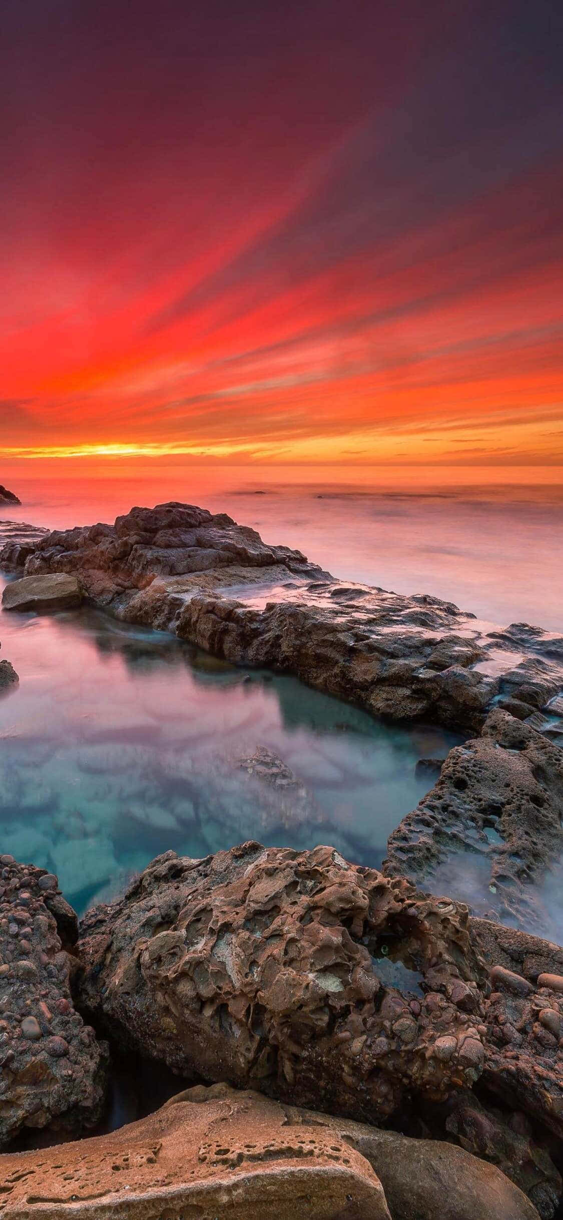 加州海岸日出 壁紙 — 熾熱的夕陽在崎嶇的海岸岩石上方將天空染成紅色、橙色和紫色，下方是碧綠色的海水。