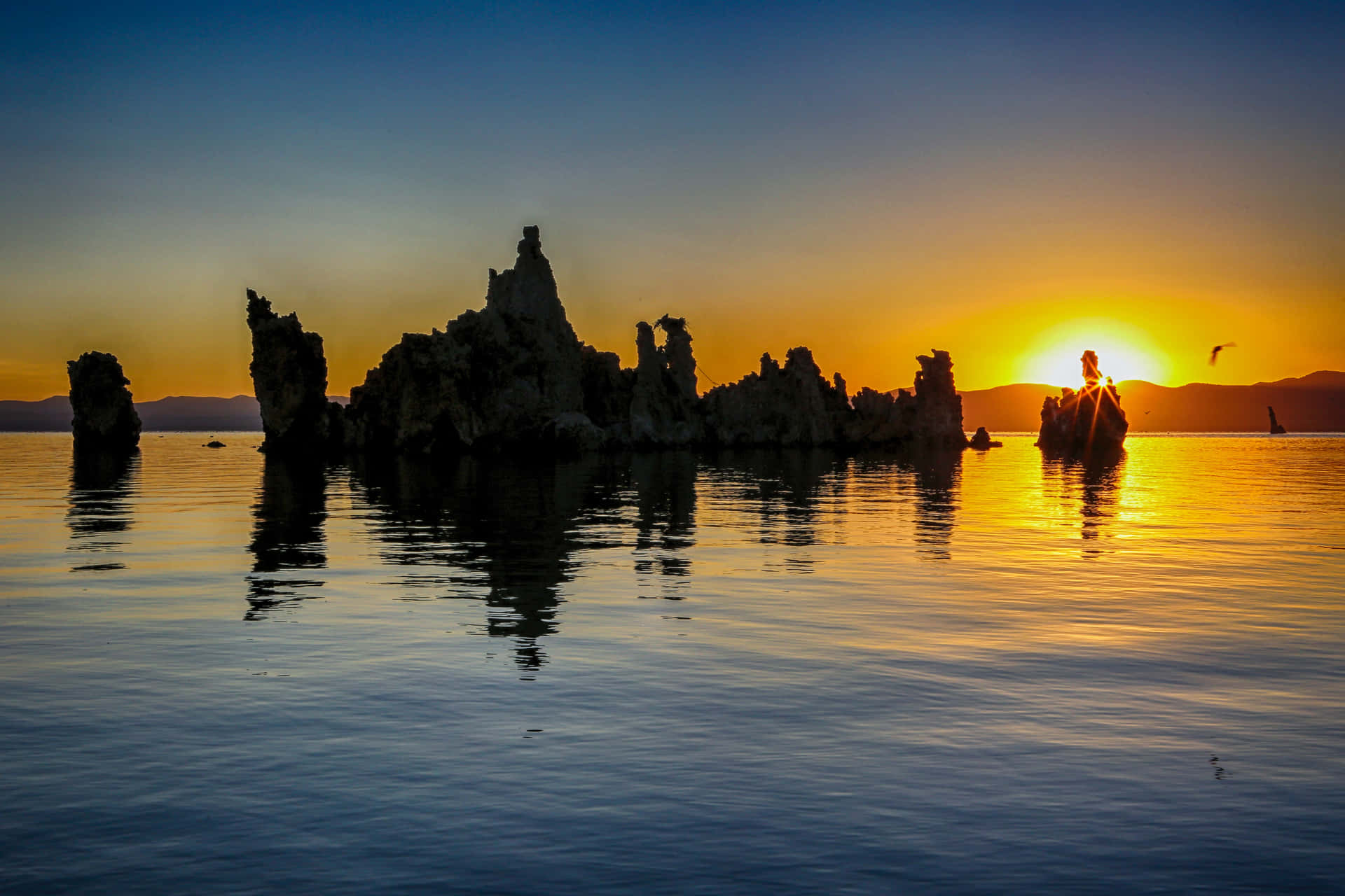 凝灰岩塔在平靜的湖面上以夕陽為背景形成剪影，山巒背景和飛鳥。