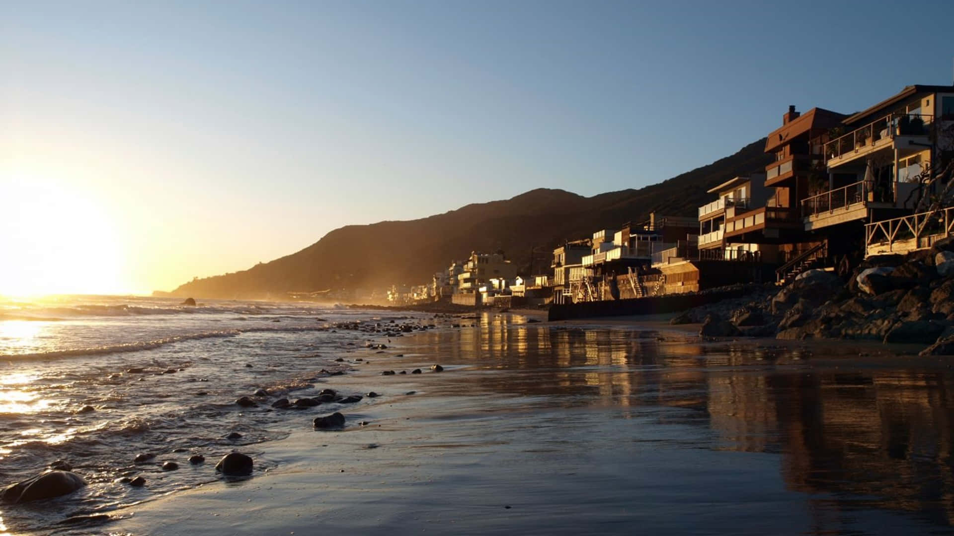 加州馬里布清晨的美麗風景 壁紙 — 夕陽以溫暖的金色光芒沐浴著沿海小鎮，海浪輕輕拍打岩石海岸。