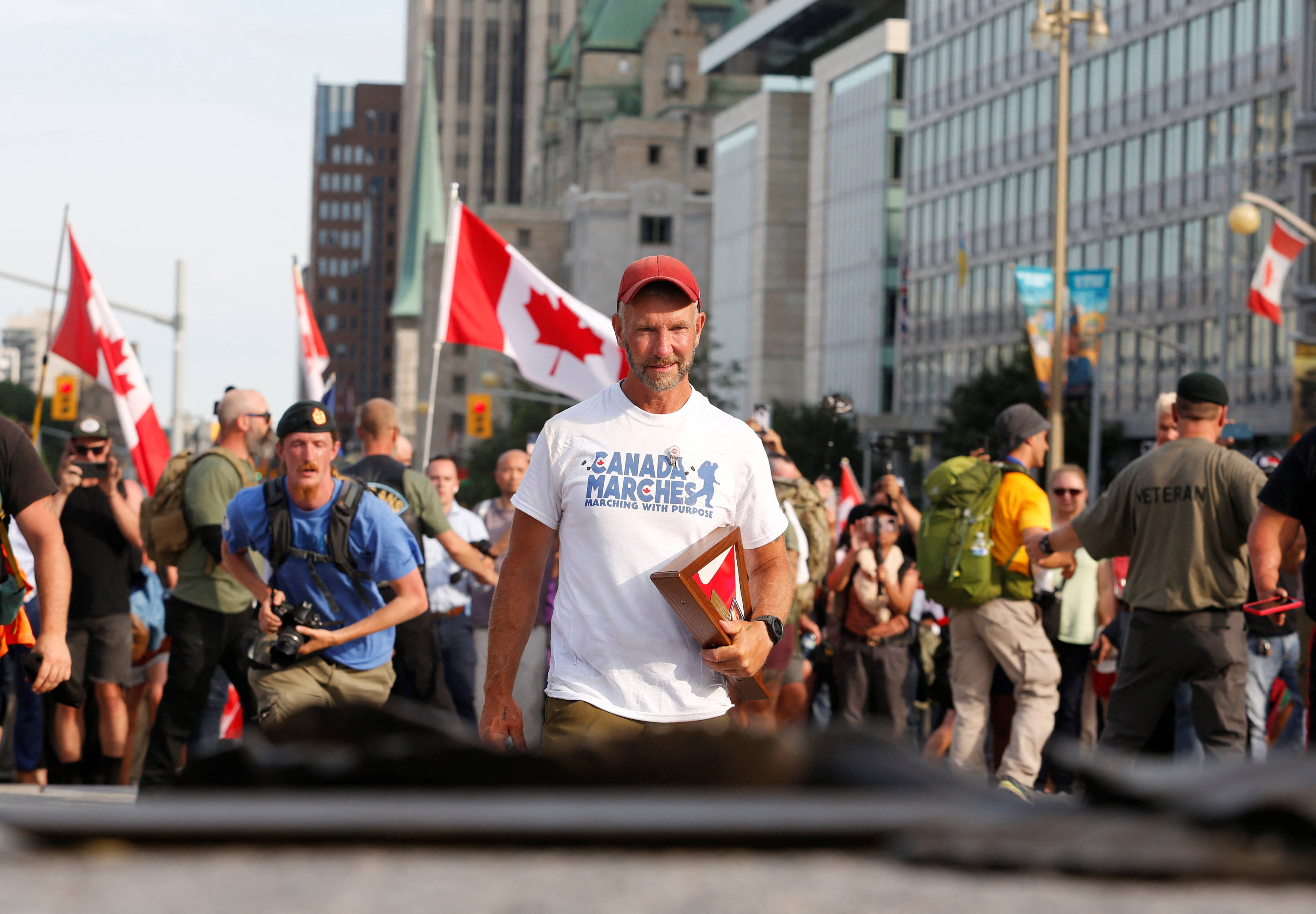 加拿大渥太華街頭活動 壁紙 — 一名戴紅色帽子和身穿「Canada Marches」T恤的男子，在日間抗議活動中手持裱框照片向前走，背景有加拿大國旗和城市建築。