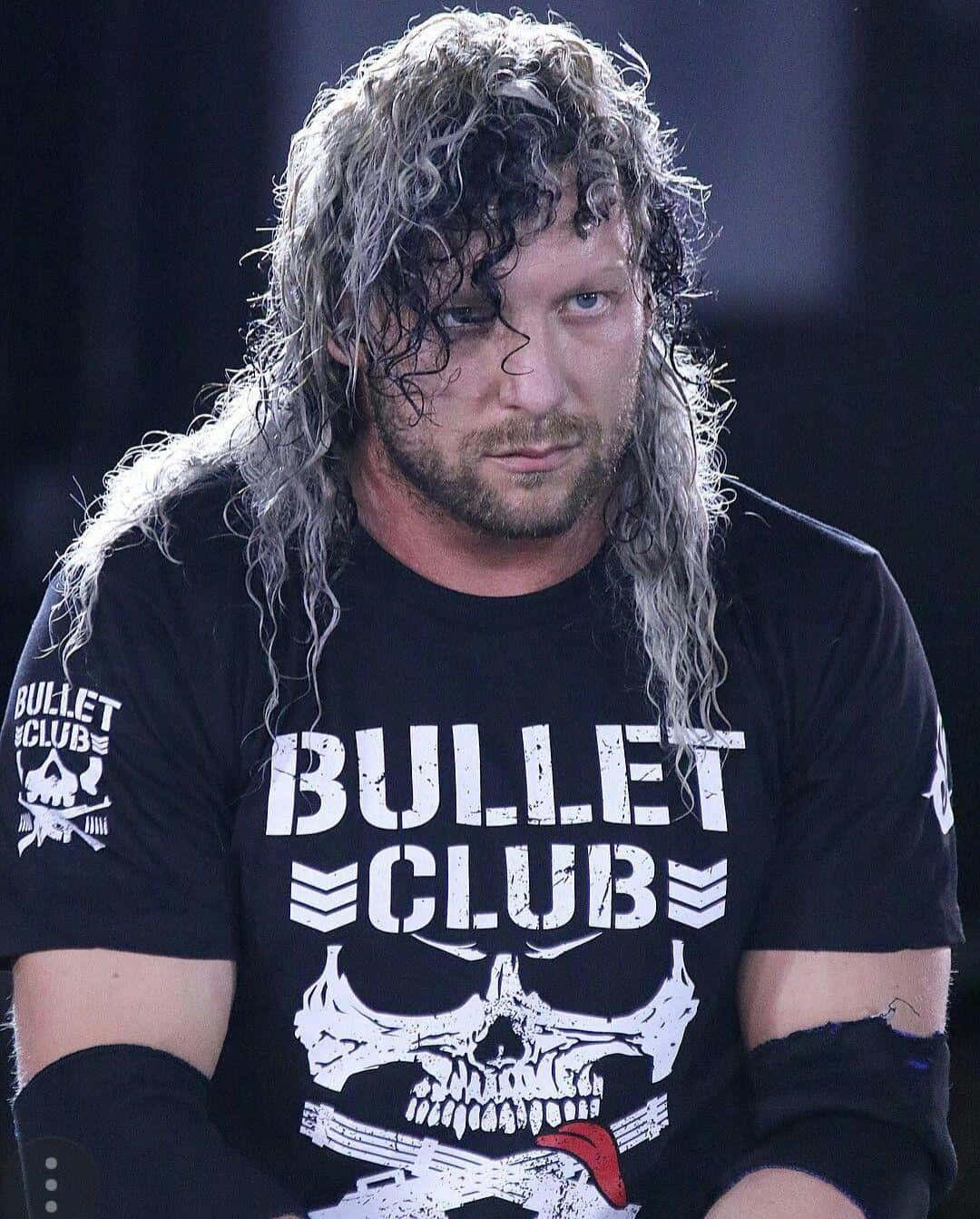留著長灰髮的摔角手，穿著印有骷髏和槍枝標誌的 Bullet Club T 恤。