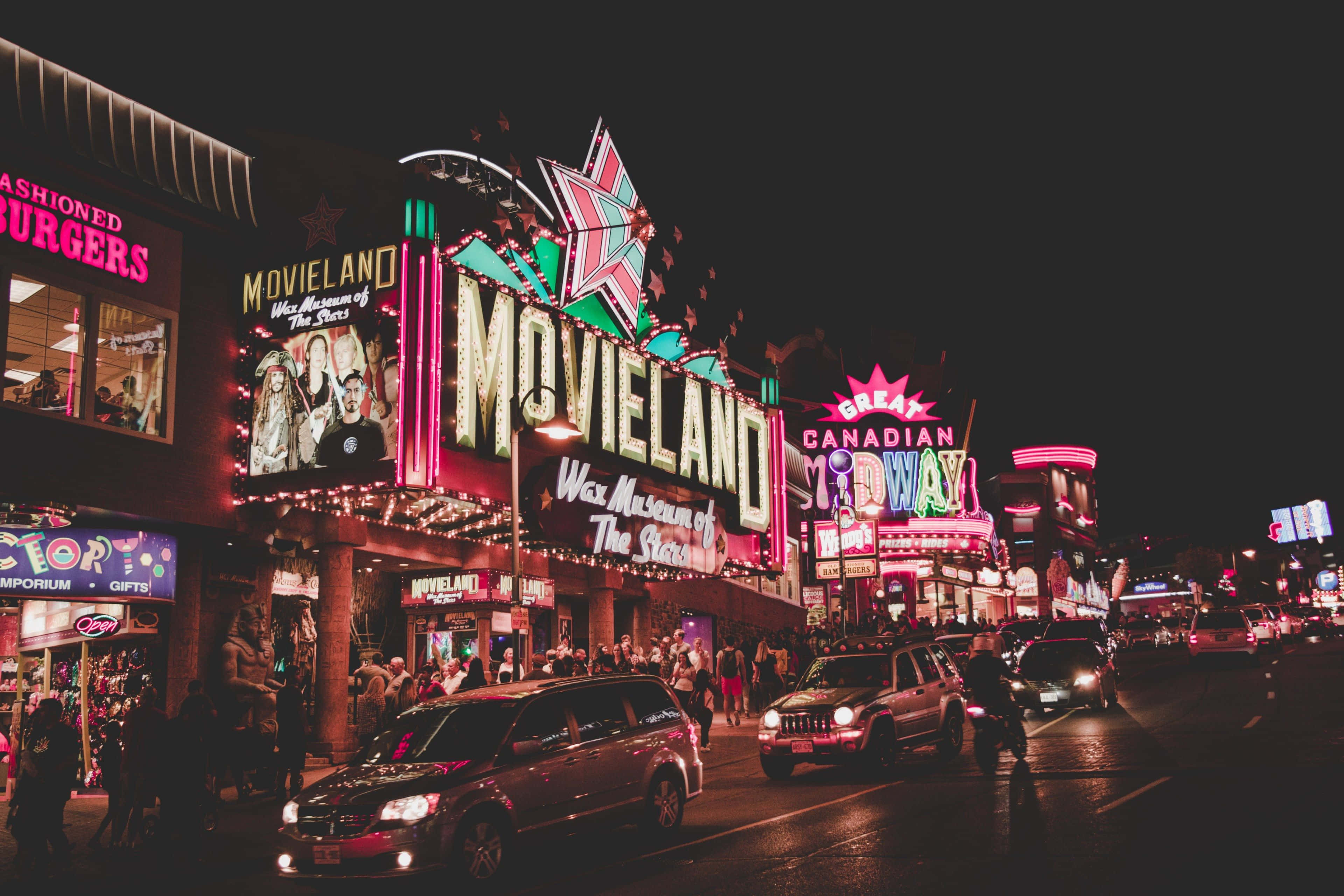 加拿大莫維爾蠟像館電影院背景 壁紙 — 鮮豔的霓虹燈照亮了Movieland蠟像館附近一條繁忙的夜間街道，有汽車和行人。