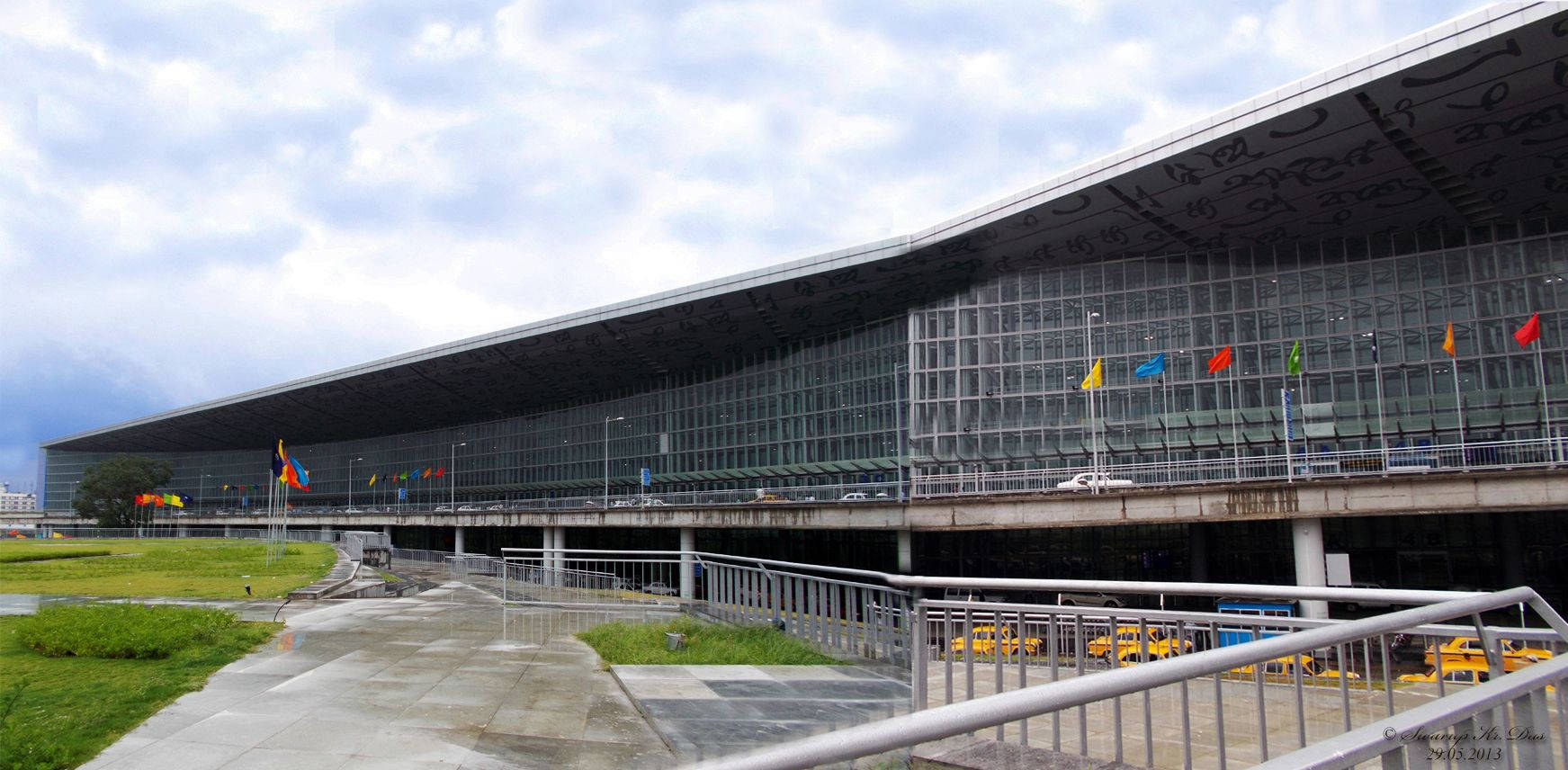 加爾各答國際機場 壁紙 — 現代機場客運大樓，玻璃幕牆和弧形屋頂，多雲的天空下，旗幟排列在行人道上。