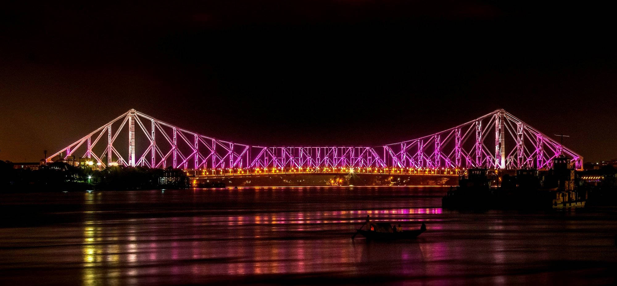 加爾各答粉紅燈光 壁紙 — 一座雄偉的鋼桁架橋在夜間發出鮮豔的紫色光芒，照亮黑暗的河流，其倒影在水面上閃爍，一艘小船在下方漂流。