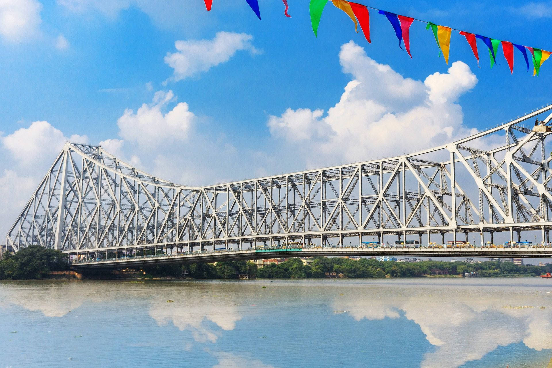 加爾各答豪拉大橋 壁紙 — 一座宏偉的鋼製桁架橋橫跨平靜的河流，上方是明亮的藍天，有蓬鬆的雲朵和色彩繽紛的三角旗。