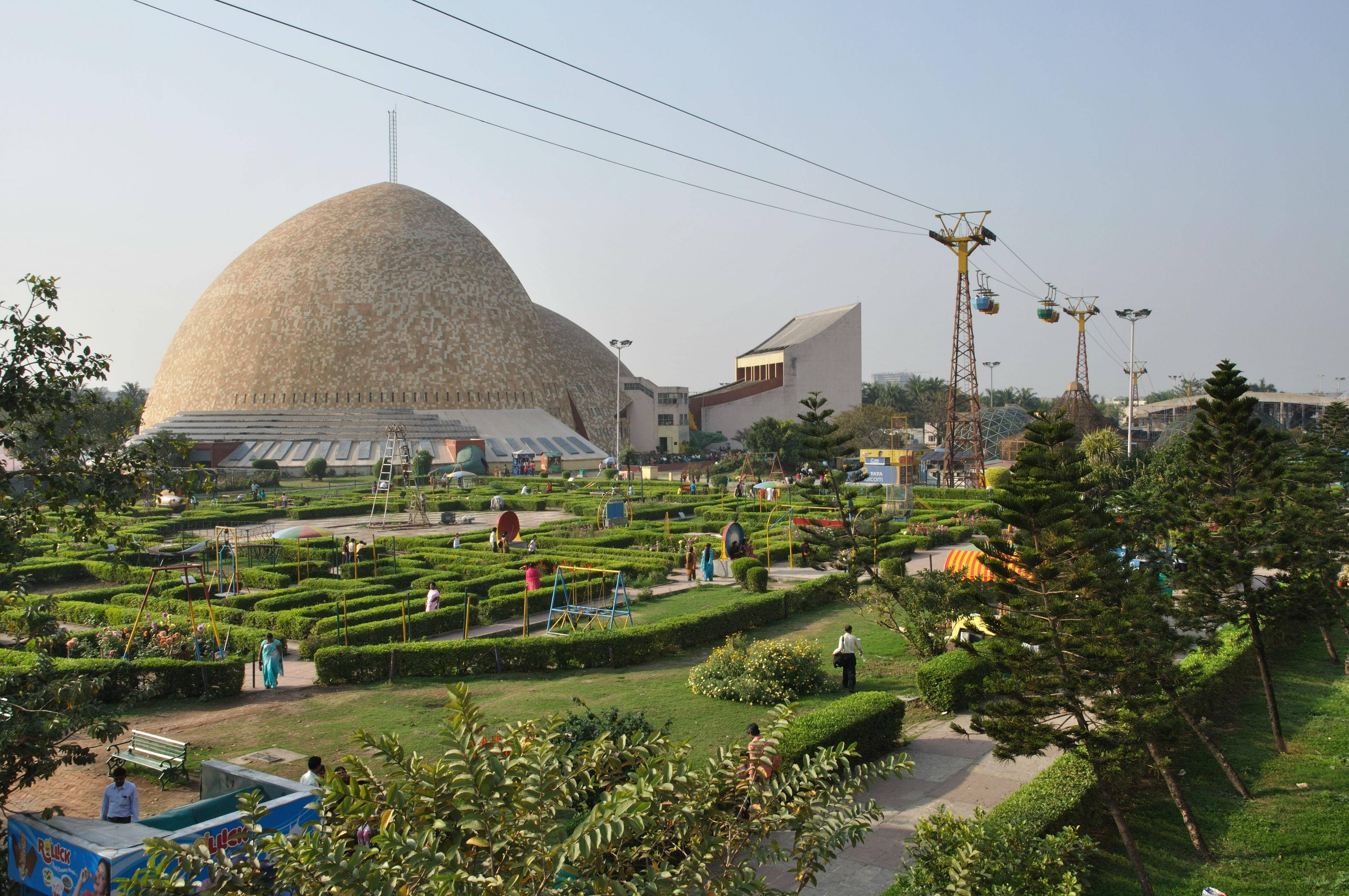 加爾各答迷宮花園 壁紙 — 一座大型圓頂建築俯瞰著一個充滿活力的花園迷宮，人們在修剪整齊的樹籬和色彩繽紛的遊樂設施之間漫步，天空晴朗。