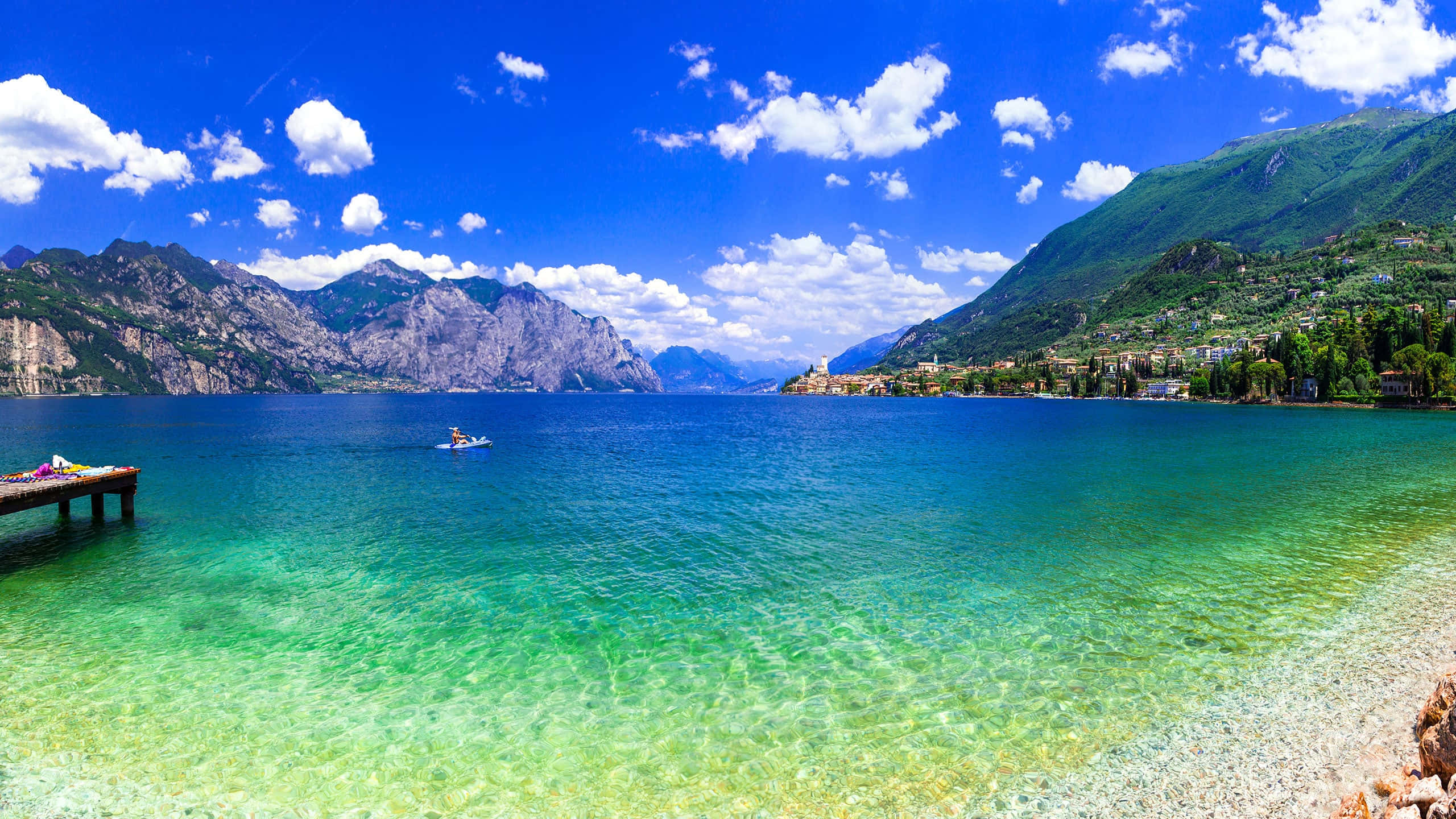 加爾達湖日間湖景 壁紙 — 一個充滿活力的藍綠色湖泊，坐落在鬱鬱蔥蔥的綠色山脈之間，上方是明亮的藍天和蓬鬆的白雲。