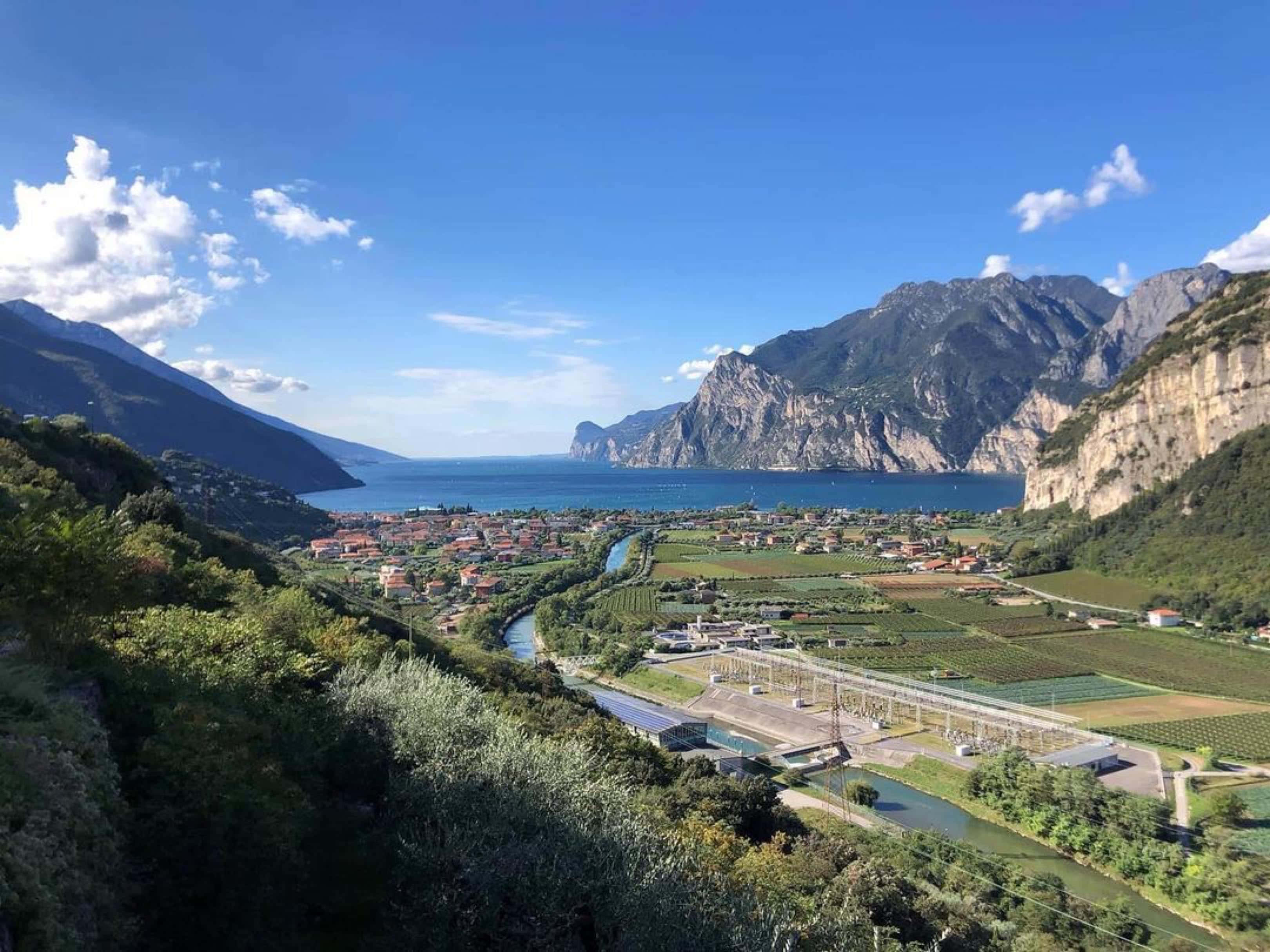 加爾達湖畔托爾博萊山景 壁紙 — 一個充滿活力的山谷俯瞰著一個深藍色的湖泊，湖泊坐落在高聳的綠色山脈之間，上方是明亮的藍天和蓬鬆的白雲。