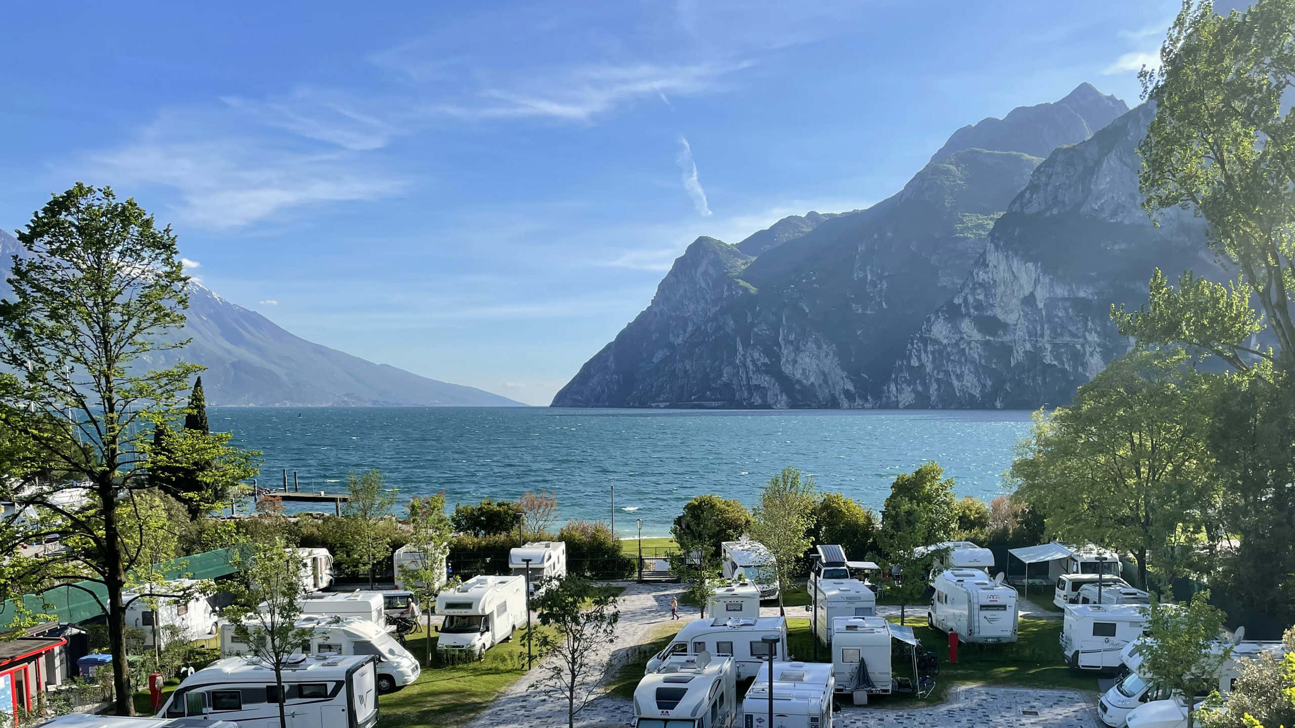 加爾達湖畔馬羅阿迪露營地 壁紙 — 白色露營車停在草地，靠近一個碧綠色的湖泊，陡峭的綠色山脈在清澈的藍天下。