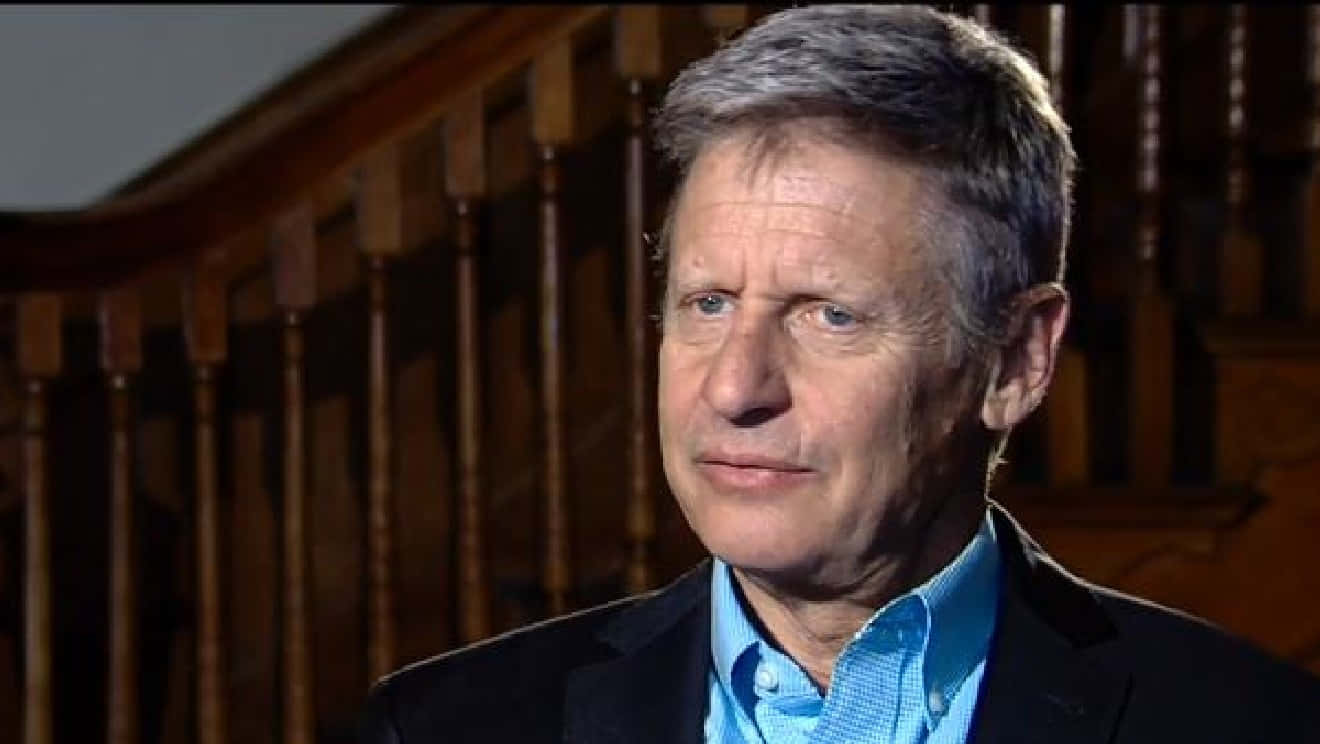一名身穿藍色襯衫和黑色西裝外套的男子，在室內靠近木樓梯站立。