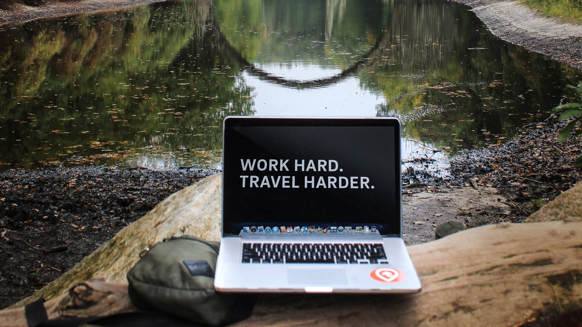 池塘旁圓木上的手提電腦，附有勵志名言 work hard travel harder