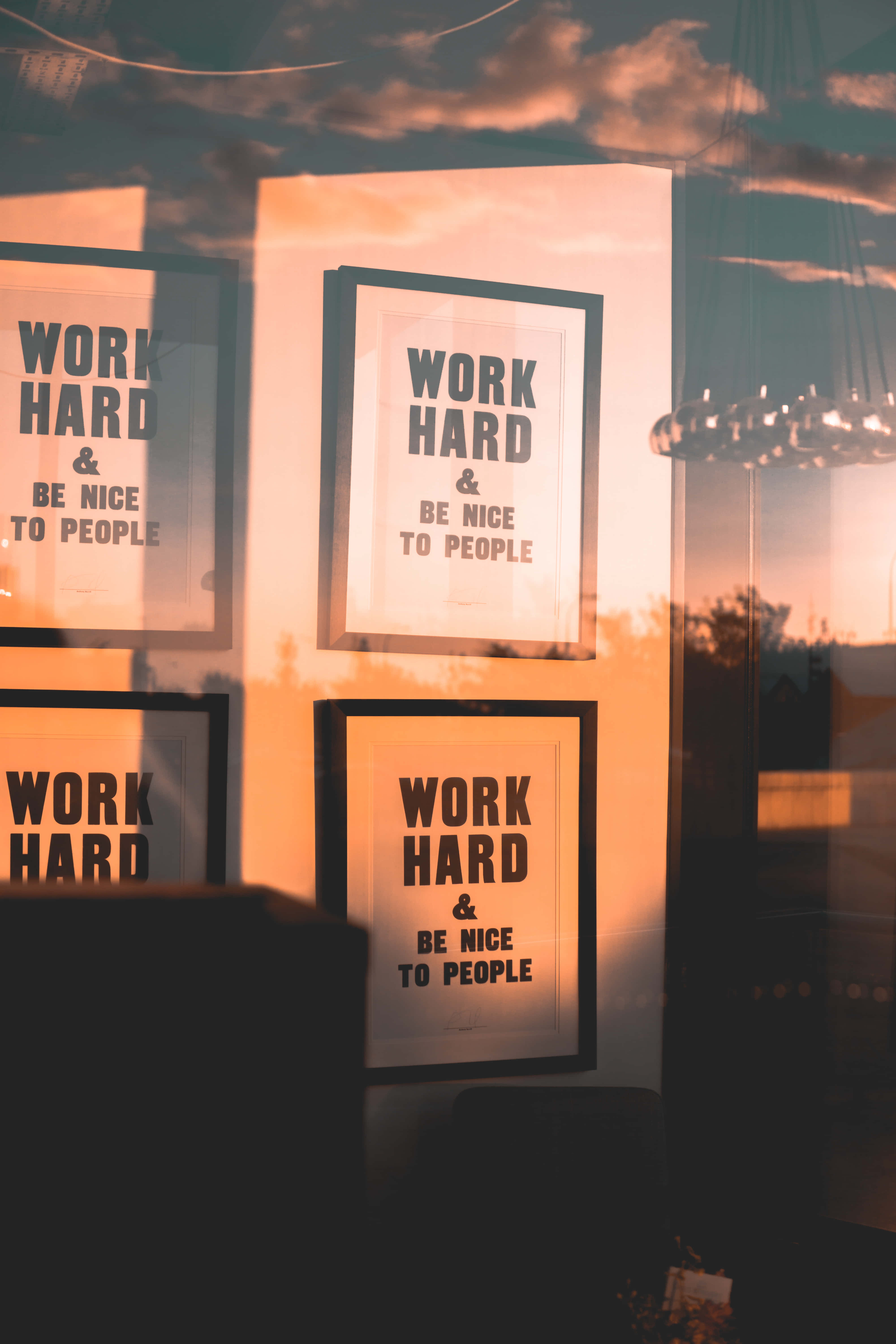 努力工作與友善待人圖片 壁紙 — 四張裱框勵志海報，上面寫著「WORK HARD & BE NICE TO PEOPLE」，在玻璃反射的日落天空襯托下溫暖地發光。