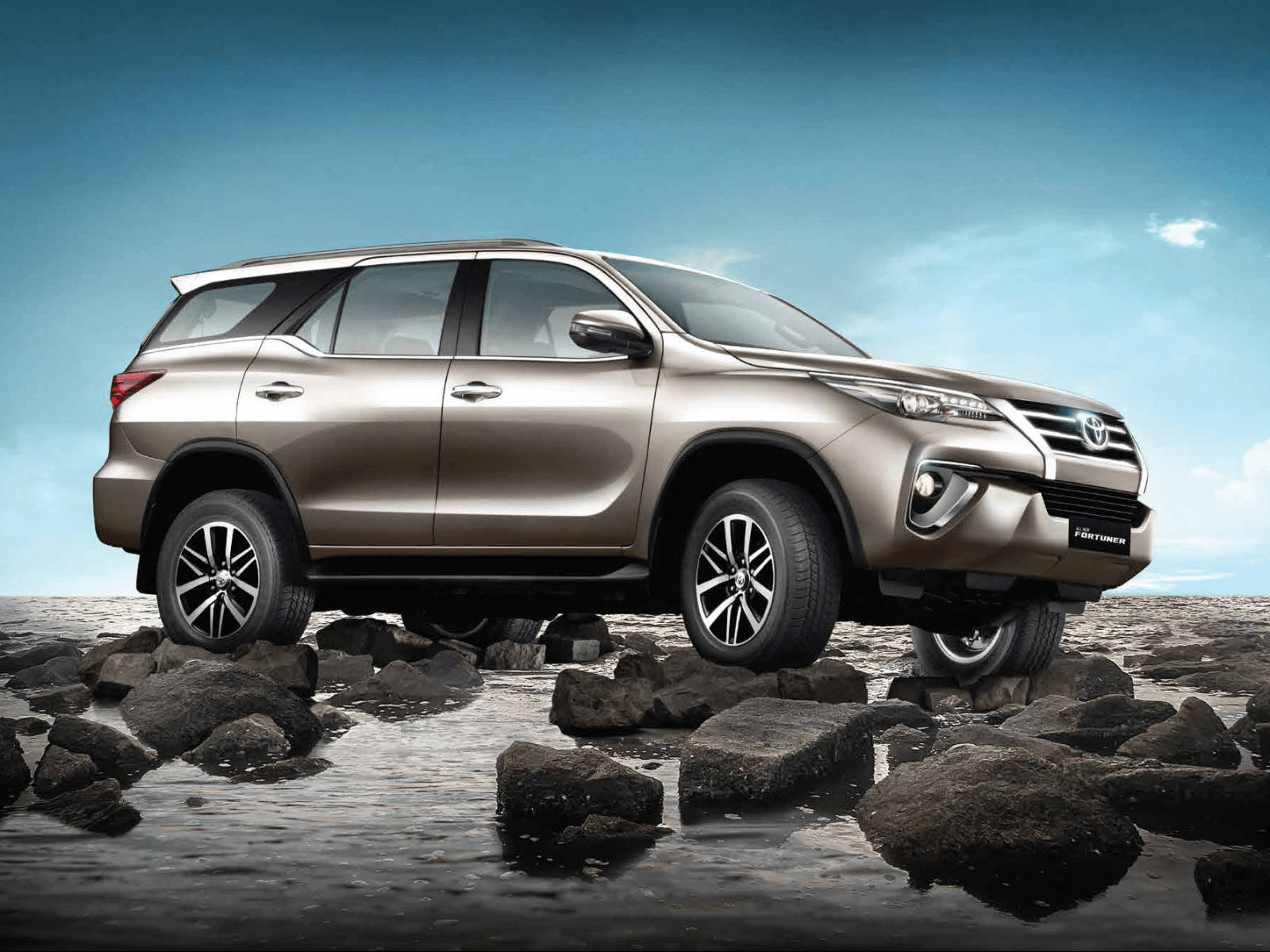 金色豐田 Fortuner SUV 在藍天下於淺水中行駛在濕岩石上。