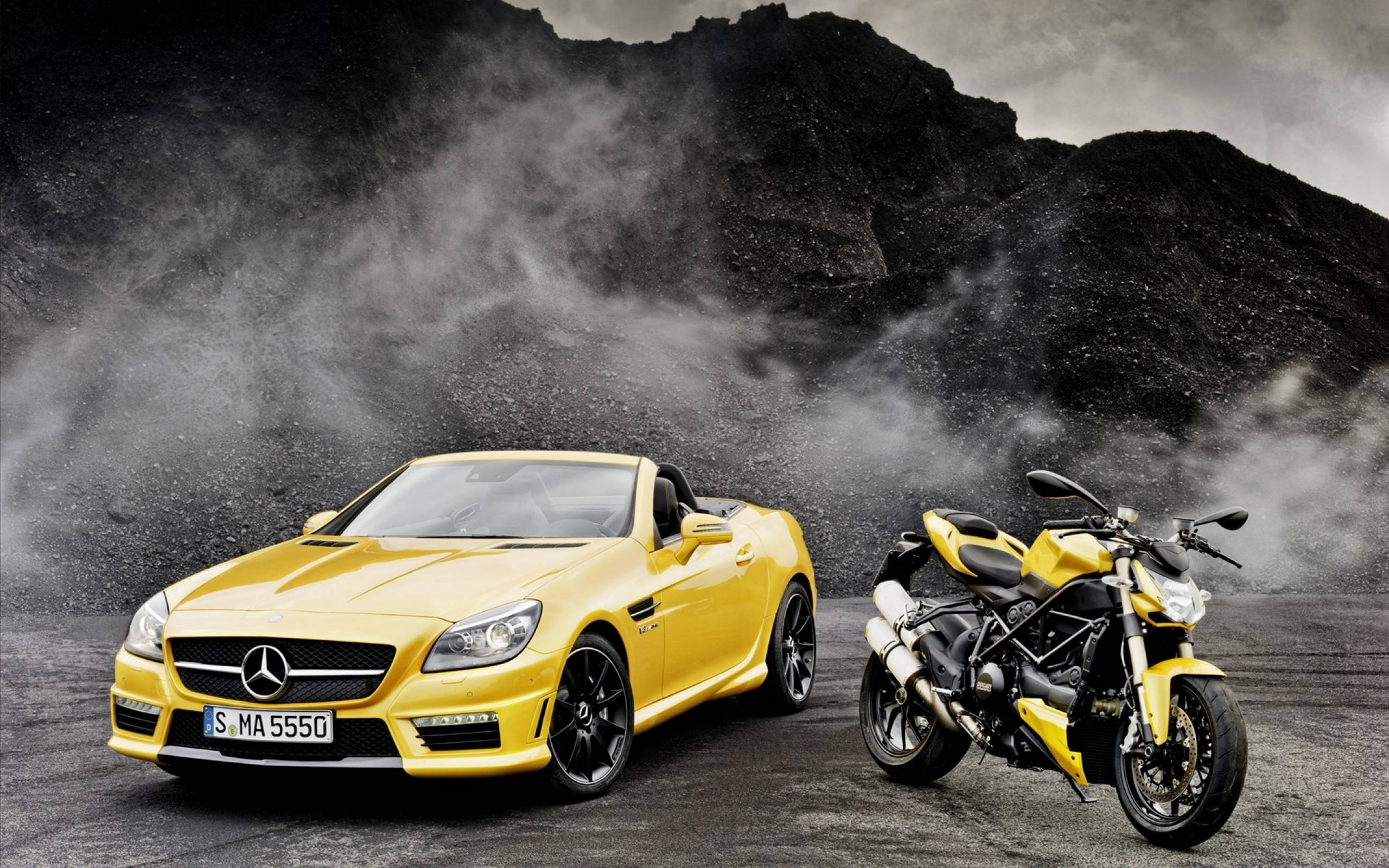 動感優雅：黃色敞篷豪華跑車 壁紙 — 一輛鮮黃色的梅賽德斯 SLK 開篷車和相配的杜卡迪電單車並排站立在塵土飛揚的火山景觀中，天空風暴密佈。