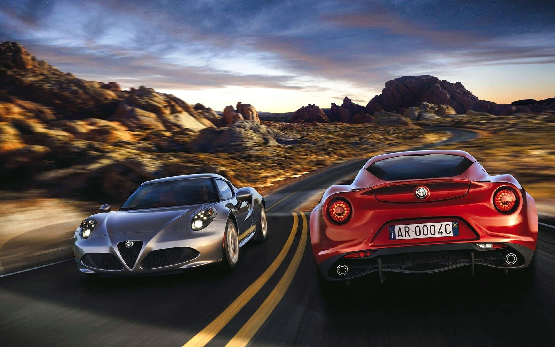 動感十足的 Alfa Romeo 4C 壁紙 — 兩部愛快羅密歐跑車在日落時分的沙漠公路上並排飛馳，背景是戲劇性的岩石地形。