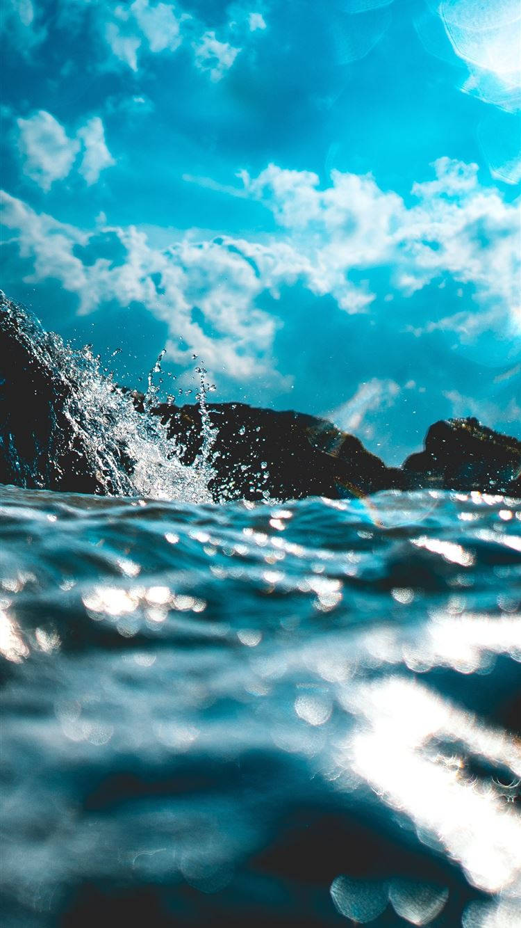 海 桌布 壁紙 — 動態的海浪拍打著深色岩石，在充滿活力的藍天和蓬鬆的白雲下。