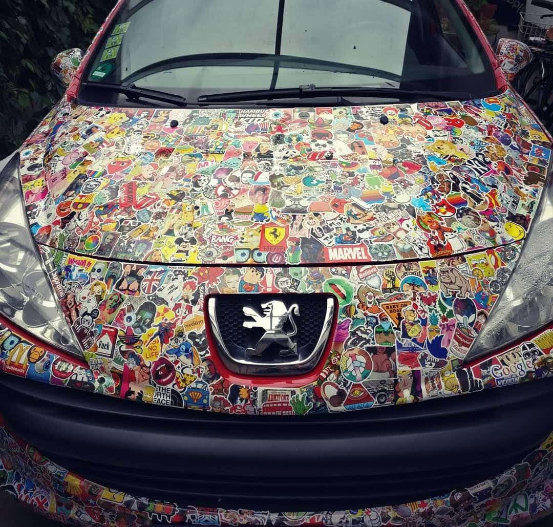 動漫與熱情驅動 壁紙 — 紅色汽車的引擎蓋上密密麻麻地貼滿了色彩繽紛的卡通和流行文化貼紙，包括Marvel和Peugeot標誌。