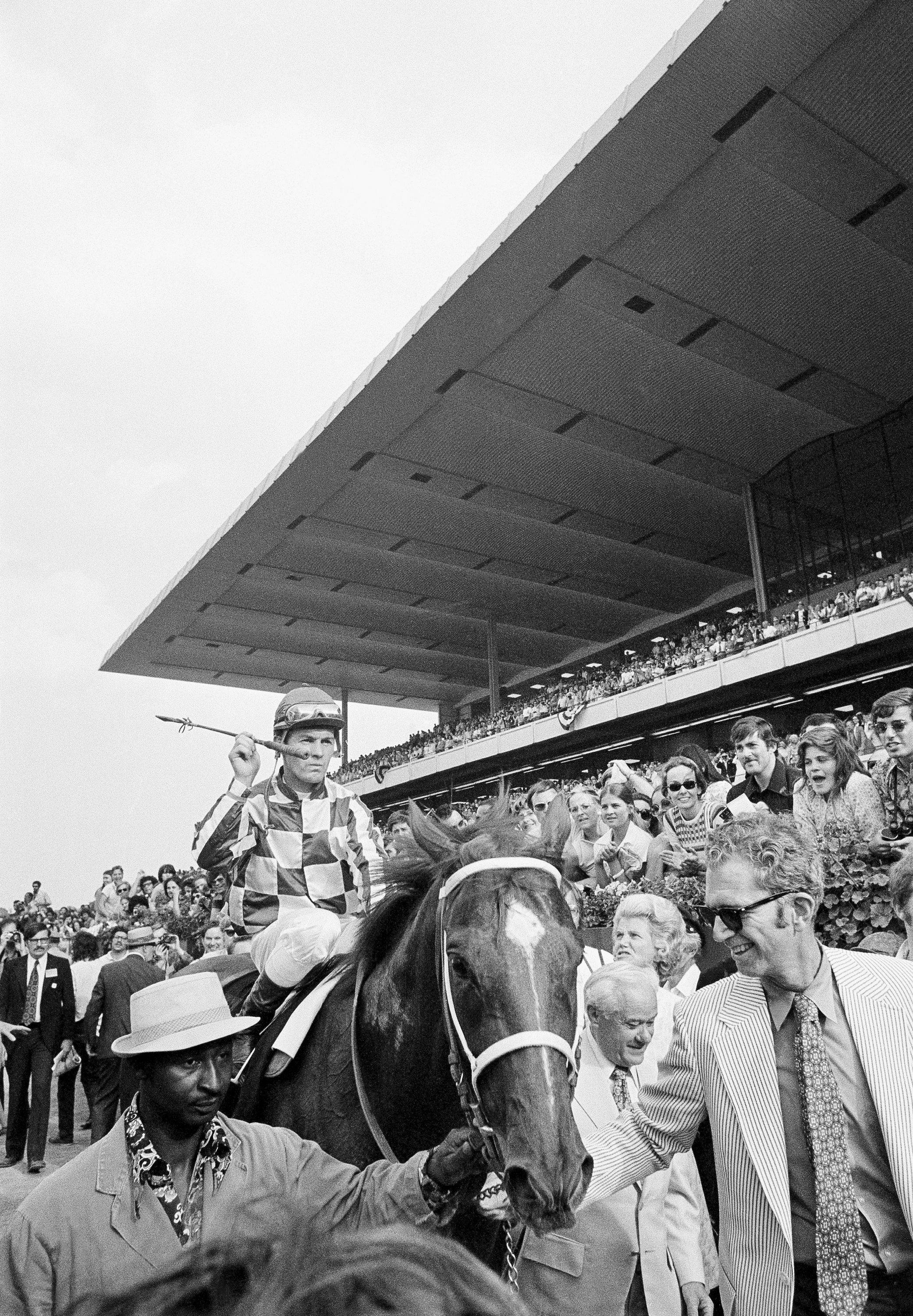 勝利時刻：Secretariat 勝出賽事 壁紙 — 一位身穿格子絲綢的騎師，在大型現代看台下的賽馬場上，騎著一匹深色馬穿過人群。