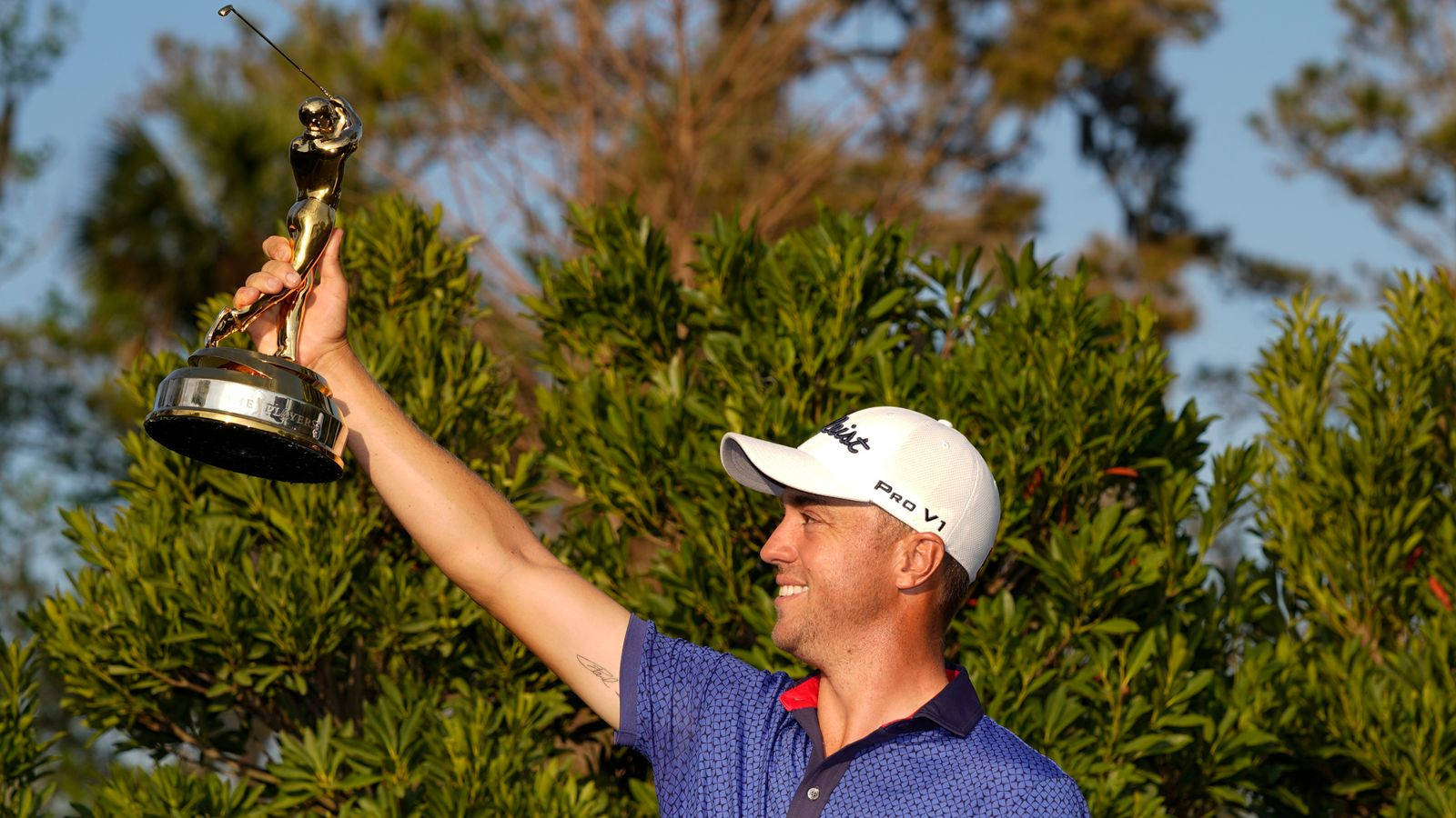 勝利的象徵：Justin Thomas 高舉獎盃 壁紙 — 一位微笑的高爾夫球手在明亮的日光下，以茂密的綠色灌木為背景，勝利地舉起閃閃發光的獎盃。