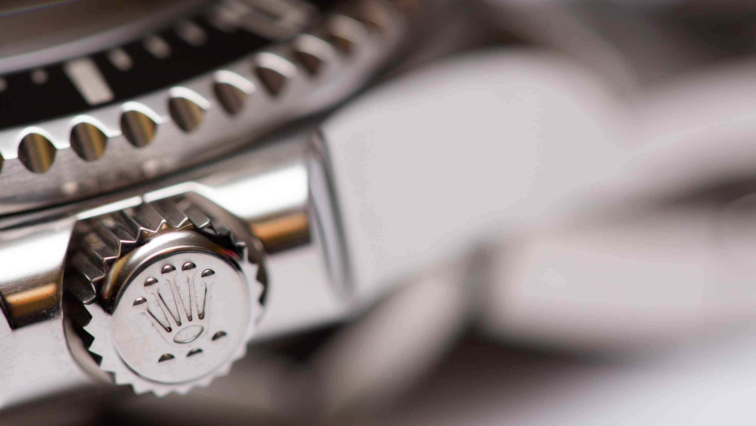 Rolex 桌布 壁紙 — 勞力士手錶錶冠特寫，帶有雕刻標誌和紋理錶圈，背景為模糊的金屬質感。