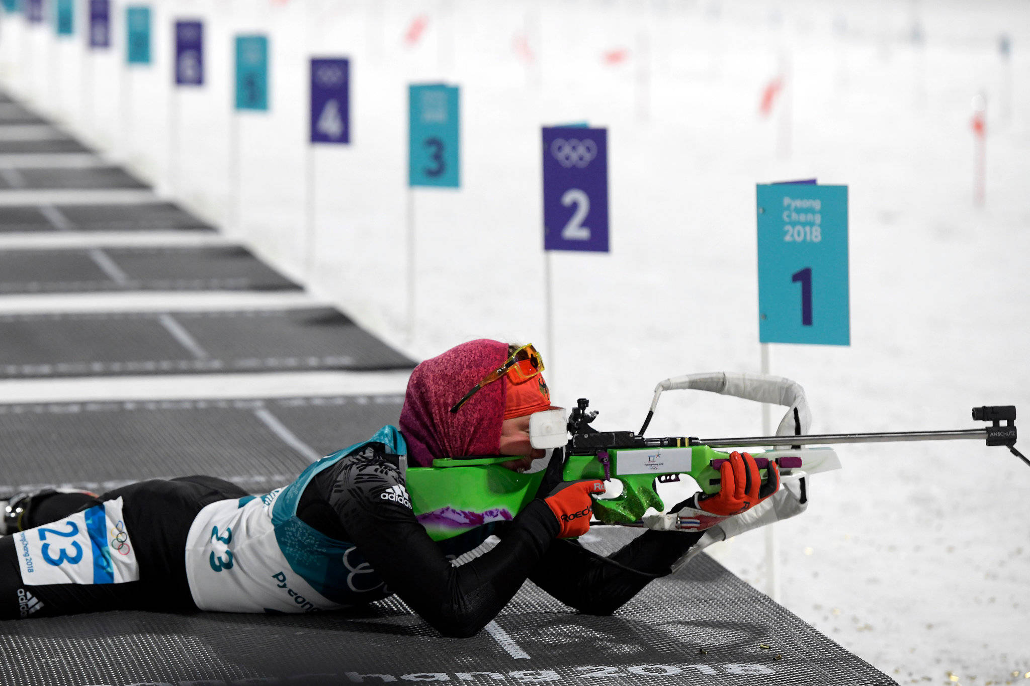 勞拉·達爾梅爾平昌冬季奧運會越野射擊場 壁紙 — 一名女性冬季兩項運動員穿著鮮豔裝備，俯臥在雪地上，用步槍瞄準帶有奧運五環和平昌2018標誌的靶標。