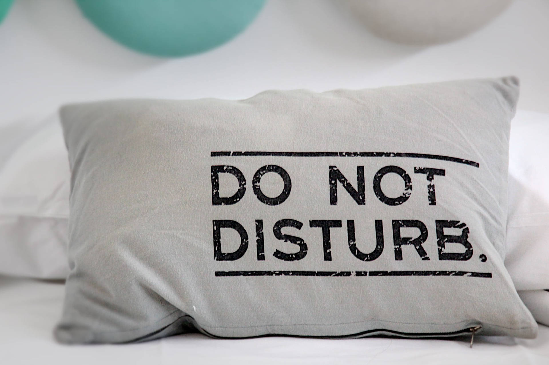灰色枕頭，上面有「DO NOT DISTURB」字樣，放在一張鋪有白色床單的床上，背景有青綠色氣球。