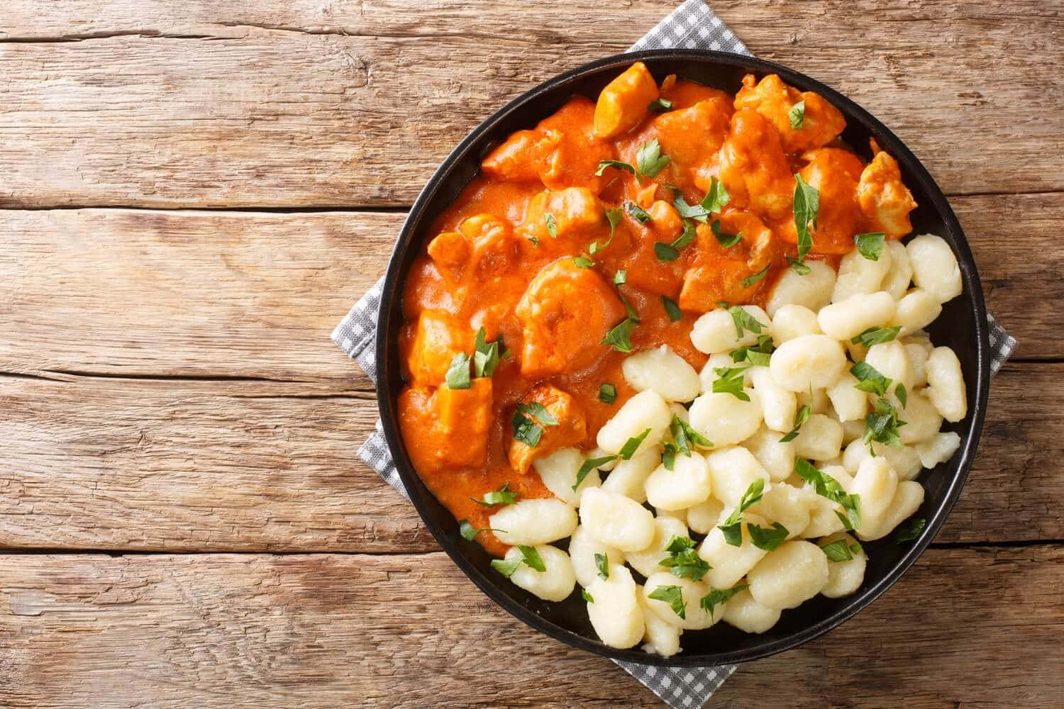 匈牙利鴨肉燉煮配義大利 gnocchi 壁紙 — 一個黑色碗盛載著奶油馬鈴薯糰子和濃郁的橙色雞肉咖喱，上面撒上新鮮綠色香草，放在質樸的木桌上。