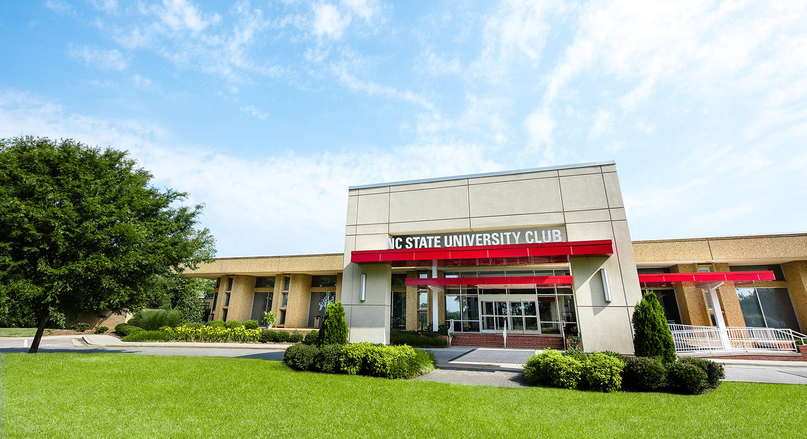 北卡羅來納州立大學俱樂部入口 壁紙 — 現代大學會所建築，設有紅色遮陽篷和綠色草坪，上方是晴朗的藍天，點綴著幾朵白雲。