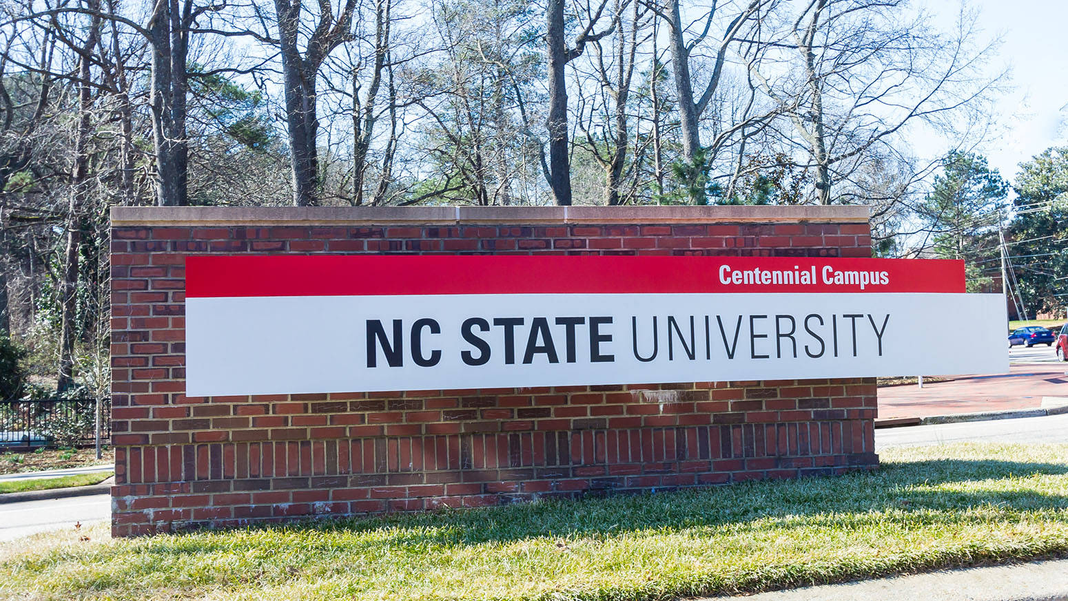 北卡羅來納州立大學校園指示牌 壁紙 — NC State University Centennial Campus 的磚製標誌矗立在草地上，背後有光禿的樹木，在明亮的日光下。