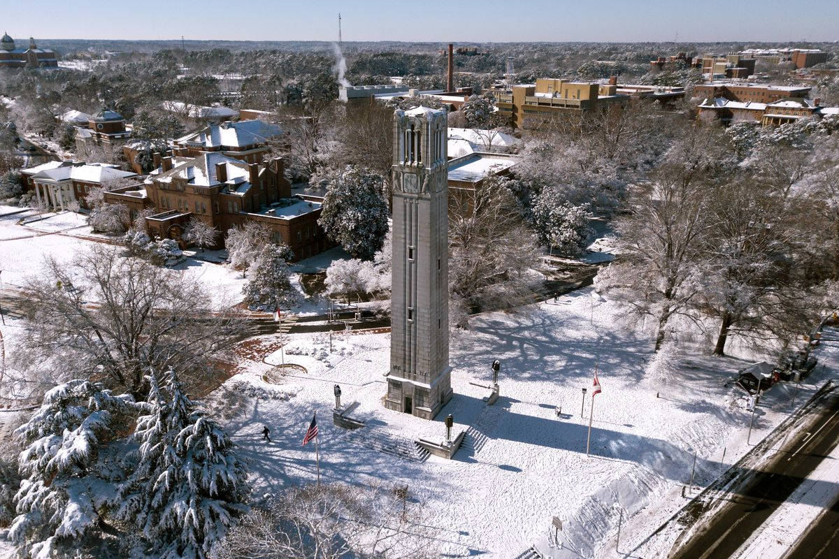 北卡羅來納州立大學雪中紀念鐘樓 壁紙 — 雪覆蓋校園的鳥瞰圖，一座高大的石造鐘樓被霜凍樹木和歷史磚砌建築環繞，在清澈藍天下。