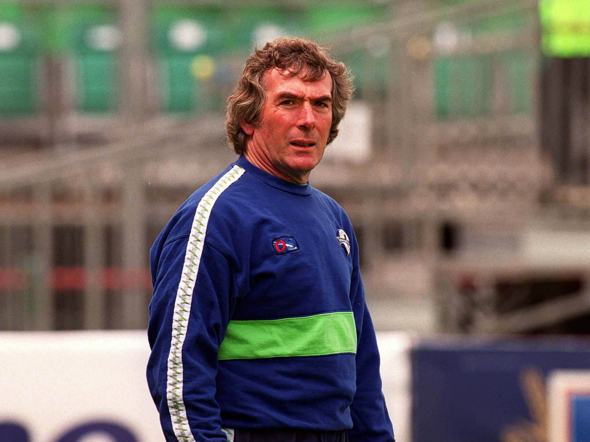 北愛爾蘭足球教練 Pat Jennings 於都柏林蘭斯當路 壁紙 — 一位專注的中年男子穿著藍綠色運動外套，站在模糊看台背景的體育場草地上。