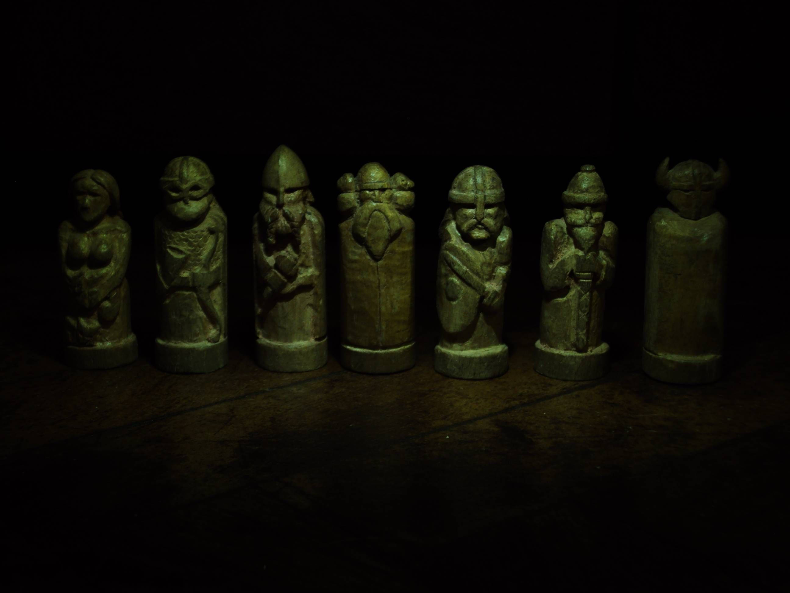 北歐神祇雕像 壁紙 — 七個古代雕刻的木製國際象棋棋子，在黑色背景前，昏暗的綠光下發光。