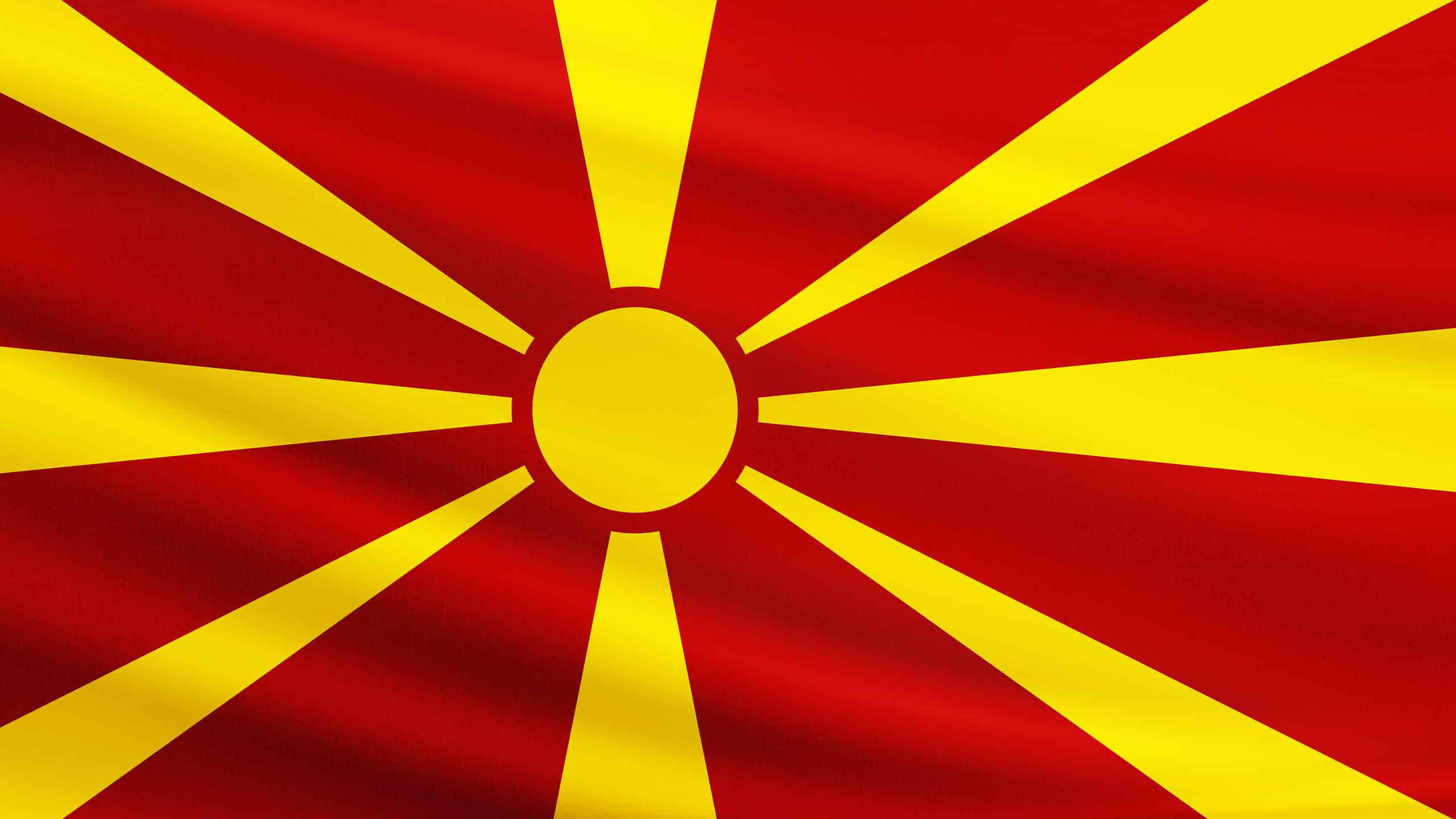 北馬其頓國旗 壁紙 — 一面鮮紅色的旗幟，帶有金色太陽爆發標誌，在晴朗的天空下動態飄揚。
