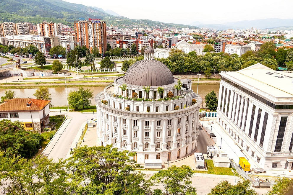 北馬其頓斯科普里 壁紙 — 一座大型白色圓頂建築，屋頂有綠植，俯瞰河流和城市景觀，四周環繞著鬱鬱蔥蔥的綠色山丘。
