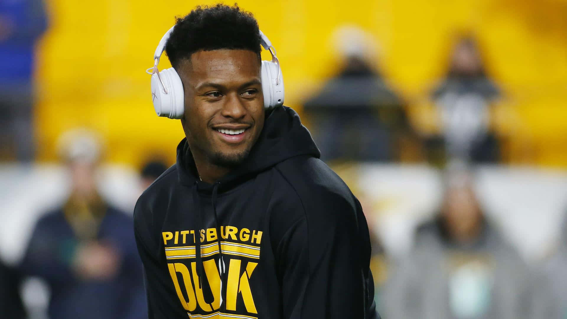 匹茲堡鋼人隊接球手 Juju Smith Schuster 壁紙 — 一位微笑的足球員穿著黑色連帽衫，上面有黃色 'PITTSBURGH DUK' 文字，戴著白色耳機，背景是模糊的體育場。