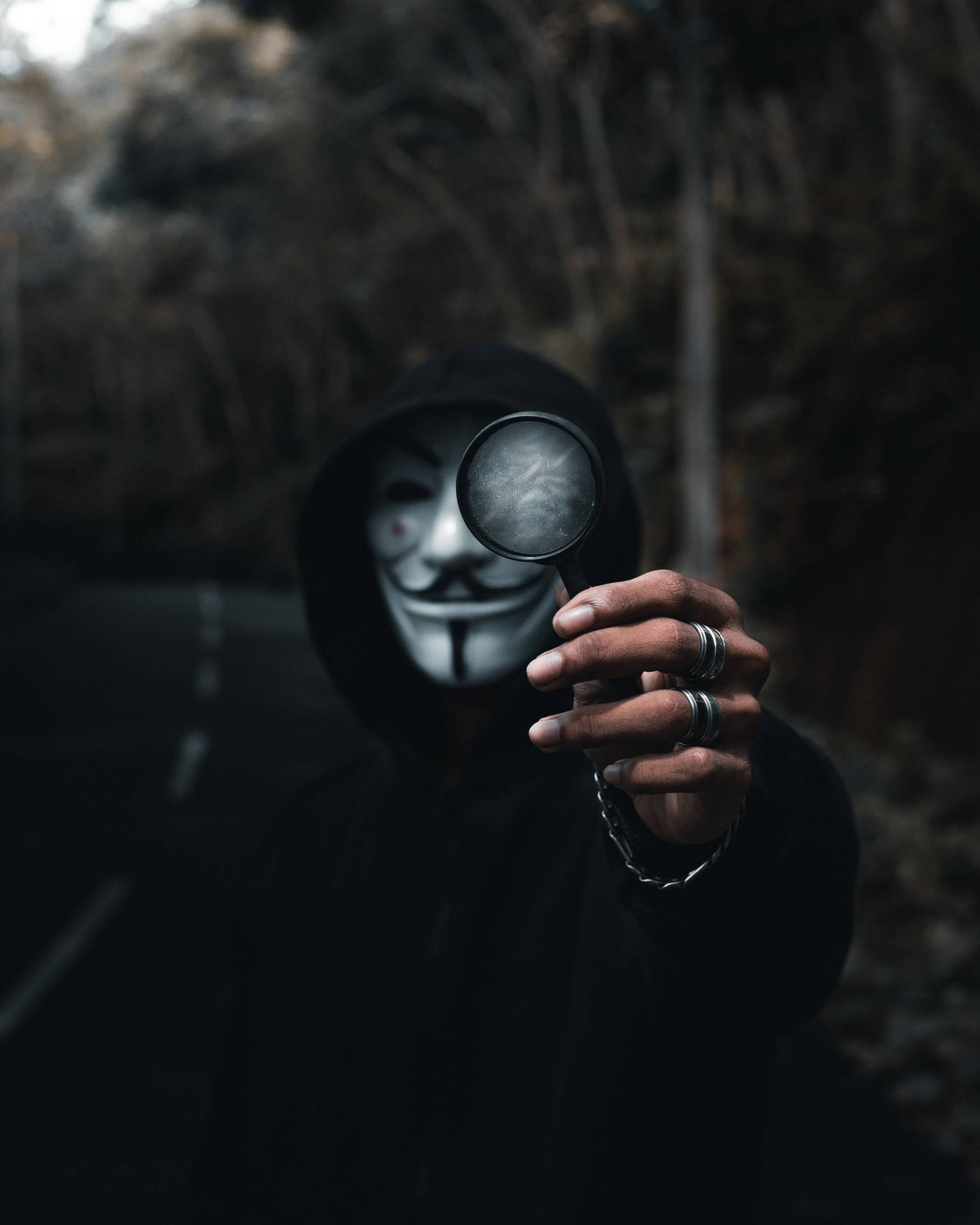 穿著黑色連帽衫和蓋伊·福克斯面具的人在戶外拿著放大鏡。