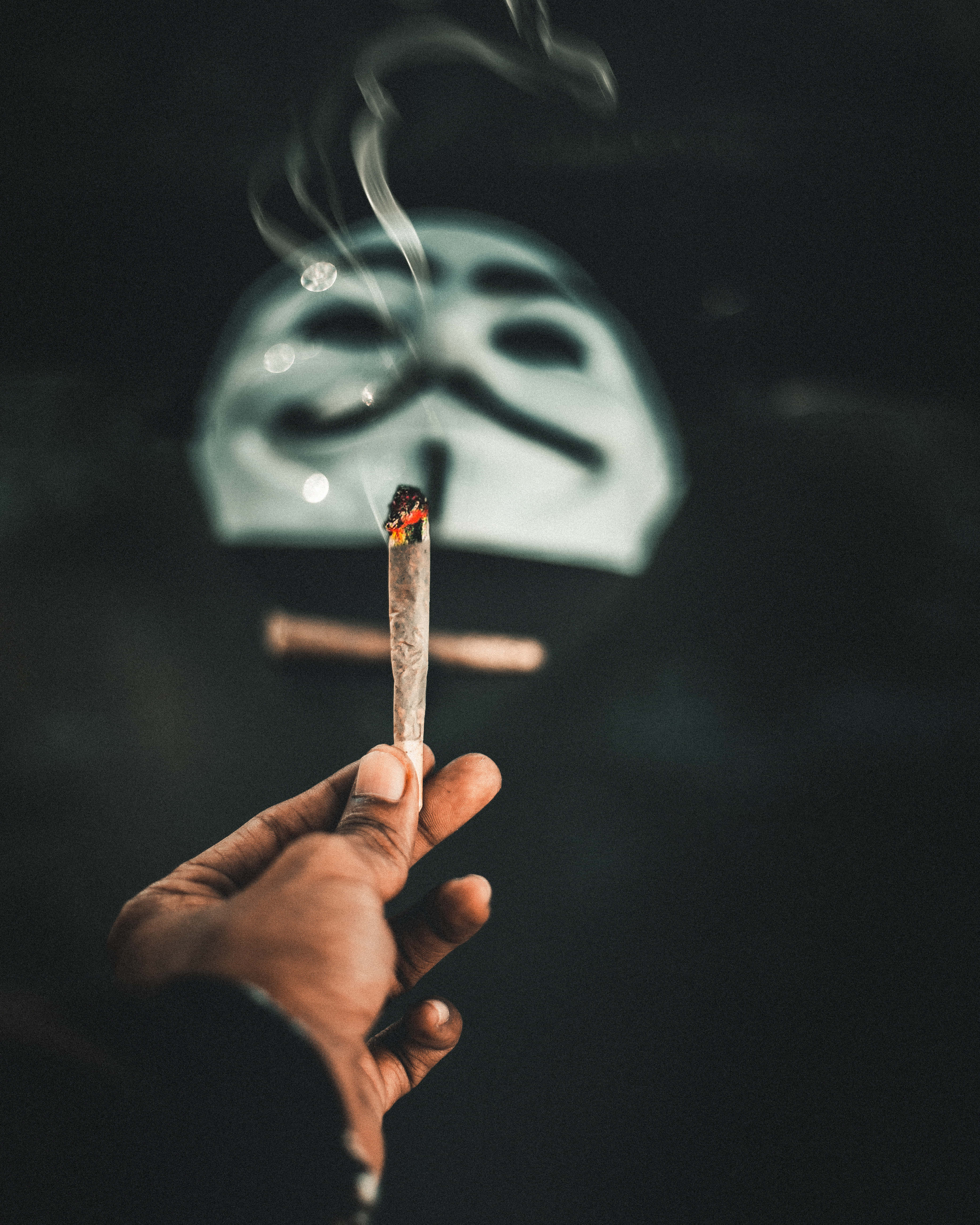 匿名與反抗：戴面具男子抽煙草 壁紙 — 一隻手拿著一支點燃的大麻煙，煙霧升起，背景是模糊的蓋伊·福克斯面具，背景黑暗。