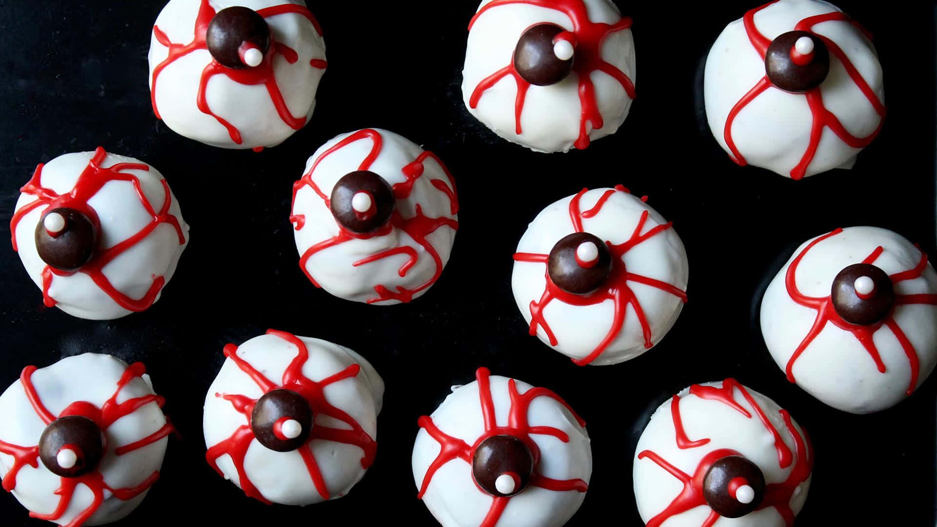 Halloween Cupcakes 桌布 壁紙 — 十二個白色糖霜點心，帶有紅色淋面和朱古力瞳孔，排列在黑色表面上。