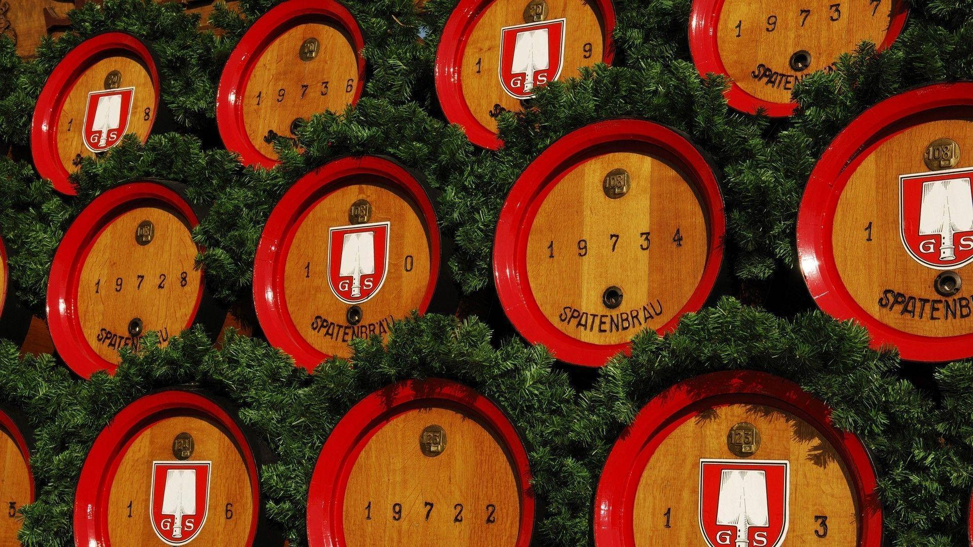 十月啤酒節啤酒桶堆疊 壁紙 — 一排排帶有紅色邊緣和 'Spatenbrau' 標誌的木製啤酒桶，藏在綠色松樹枝之間。