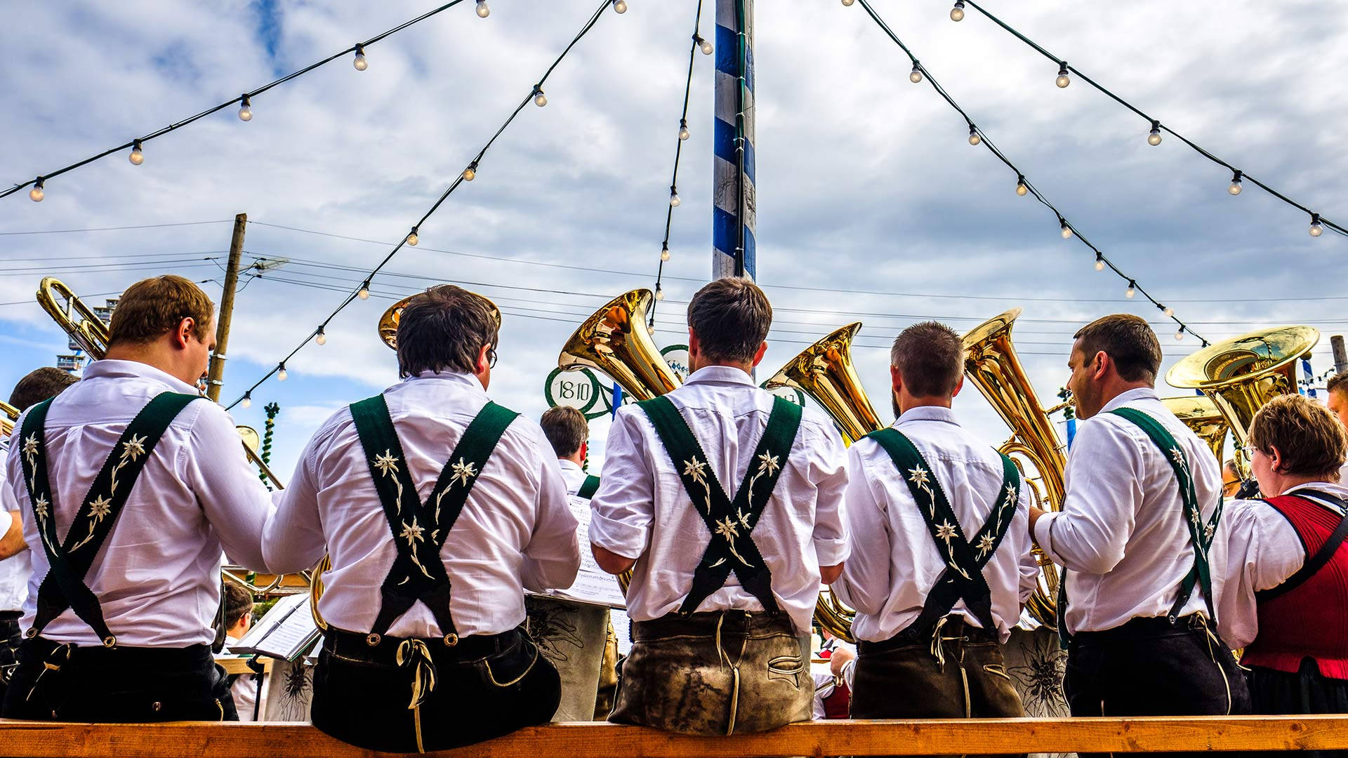 十月啤酒節管樂隊 壁紙 — 一個傳統的銅管樂隊，身穿白襯衫和綠色吊帶，在節日戶外聚會的燈串下演奏。