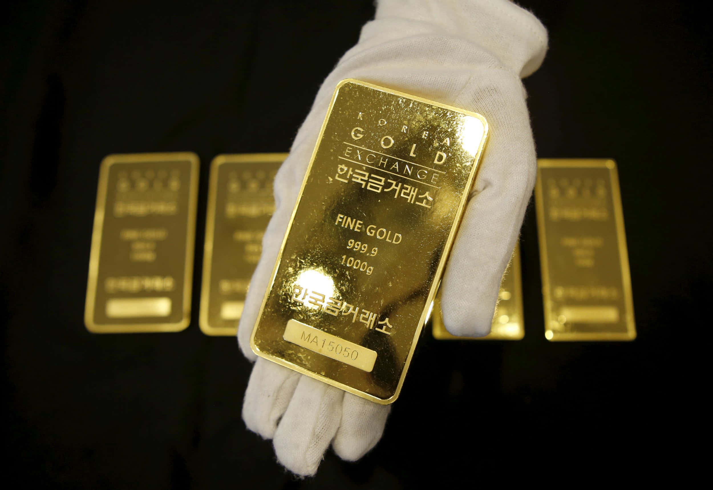 十根黃金條堆疊 壁紙 — 一隻戴著手套的手在深色背景上，與其他金條一起拿著一塊閃亮的1000g金條，上面標有Korea Gold Exchange。