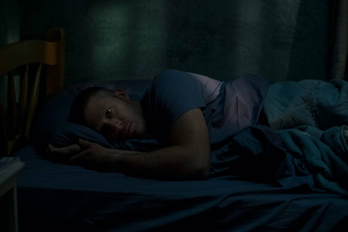 午夜彌撒瑞利·弗林臥床 壁紙 — 一個男人在昏暗的藍色燈光下躺在床上醒著，眼睛睜開，向上凝視，度過一個安靜無眠的夜晚。