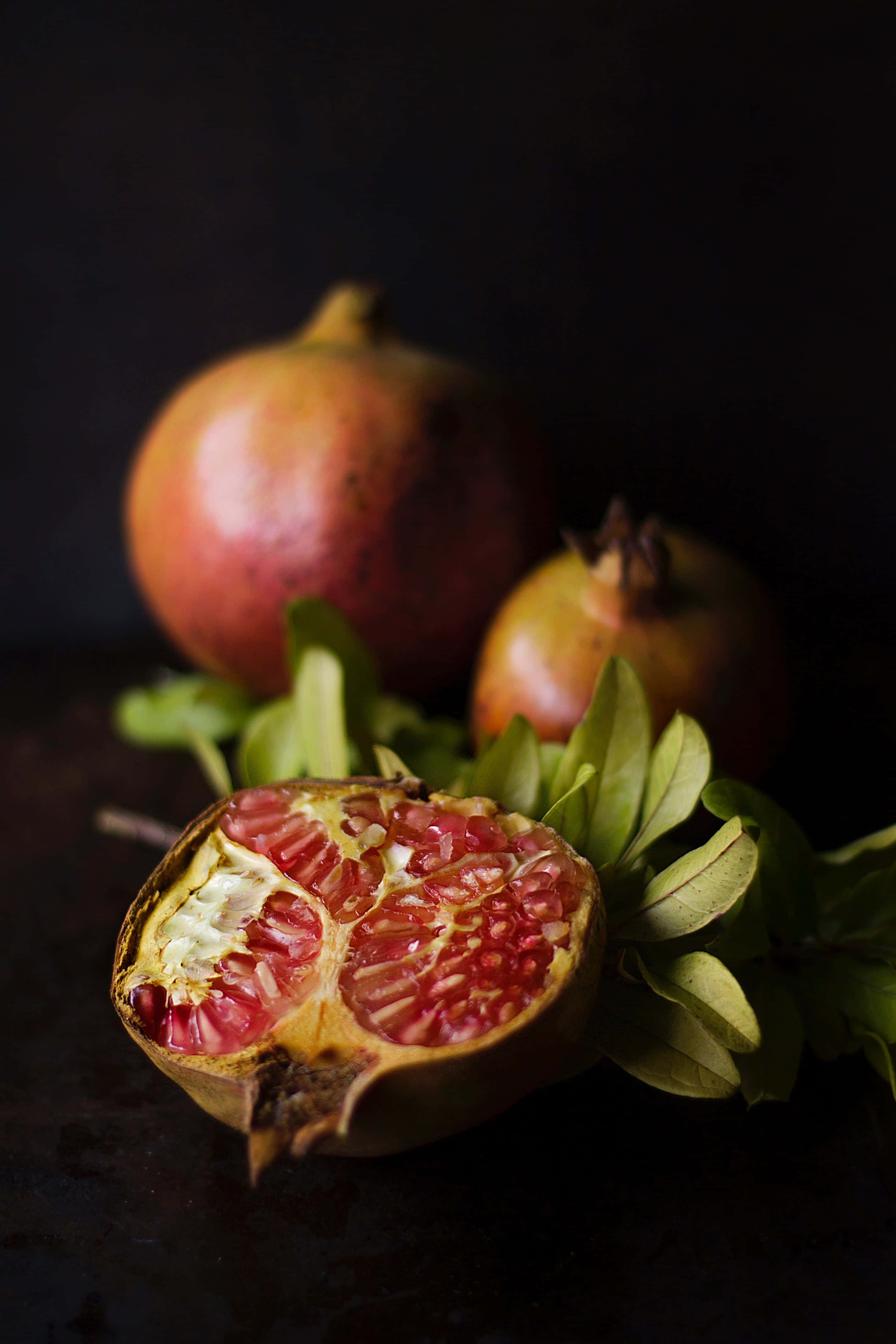 Autumn Food 桌布 壁紙 — 半個切開的石榴露出多汁的紅色籽，旁邊有完整的水果和綠葉，襯托著深色背景。