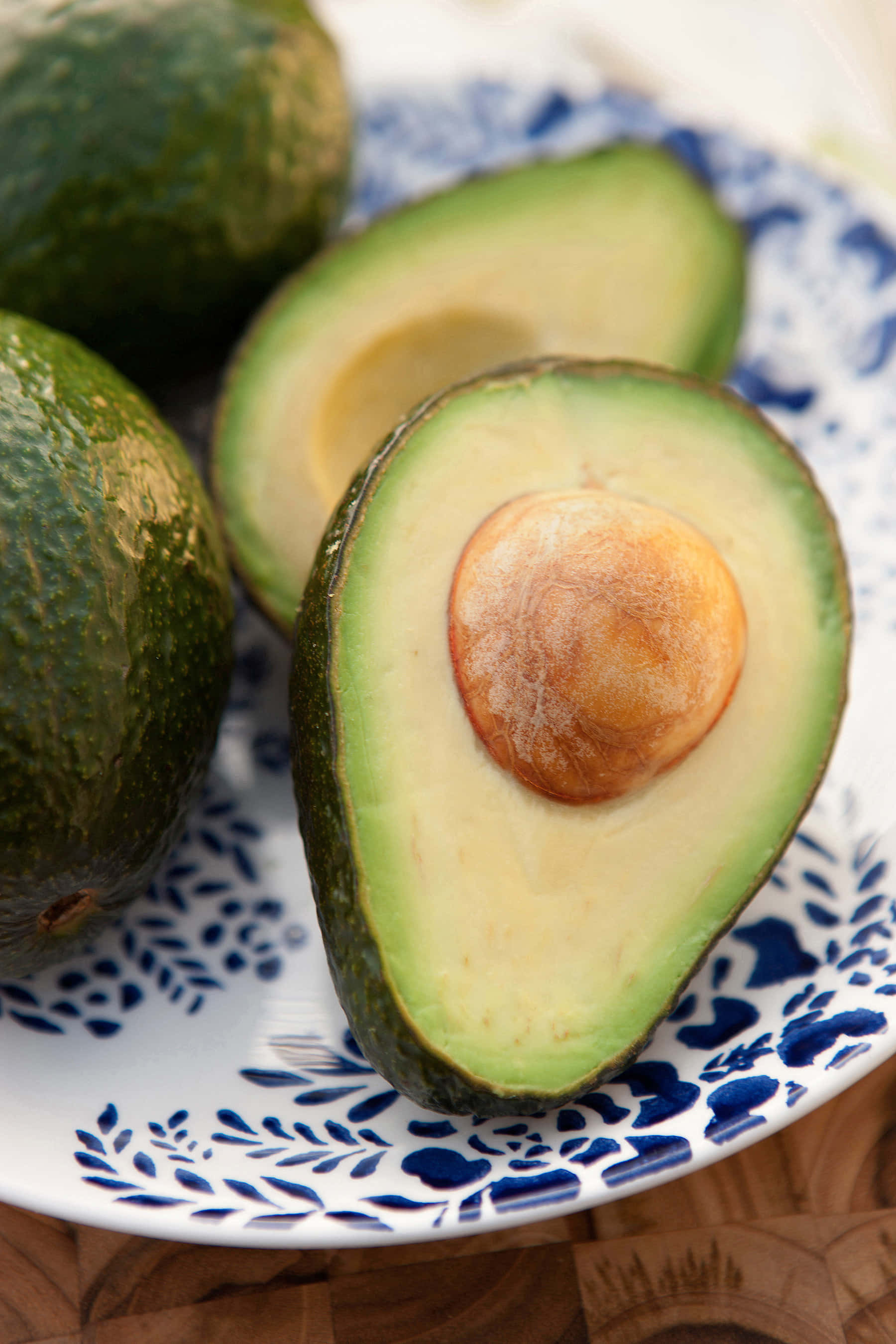 Avocado 桌布 壁紙 — 半個成熟的牛油果，帶有奶油狀的果肉和大核，放在一個藍色圖案碟上，旁邊有完整的牛油果。