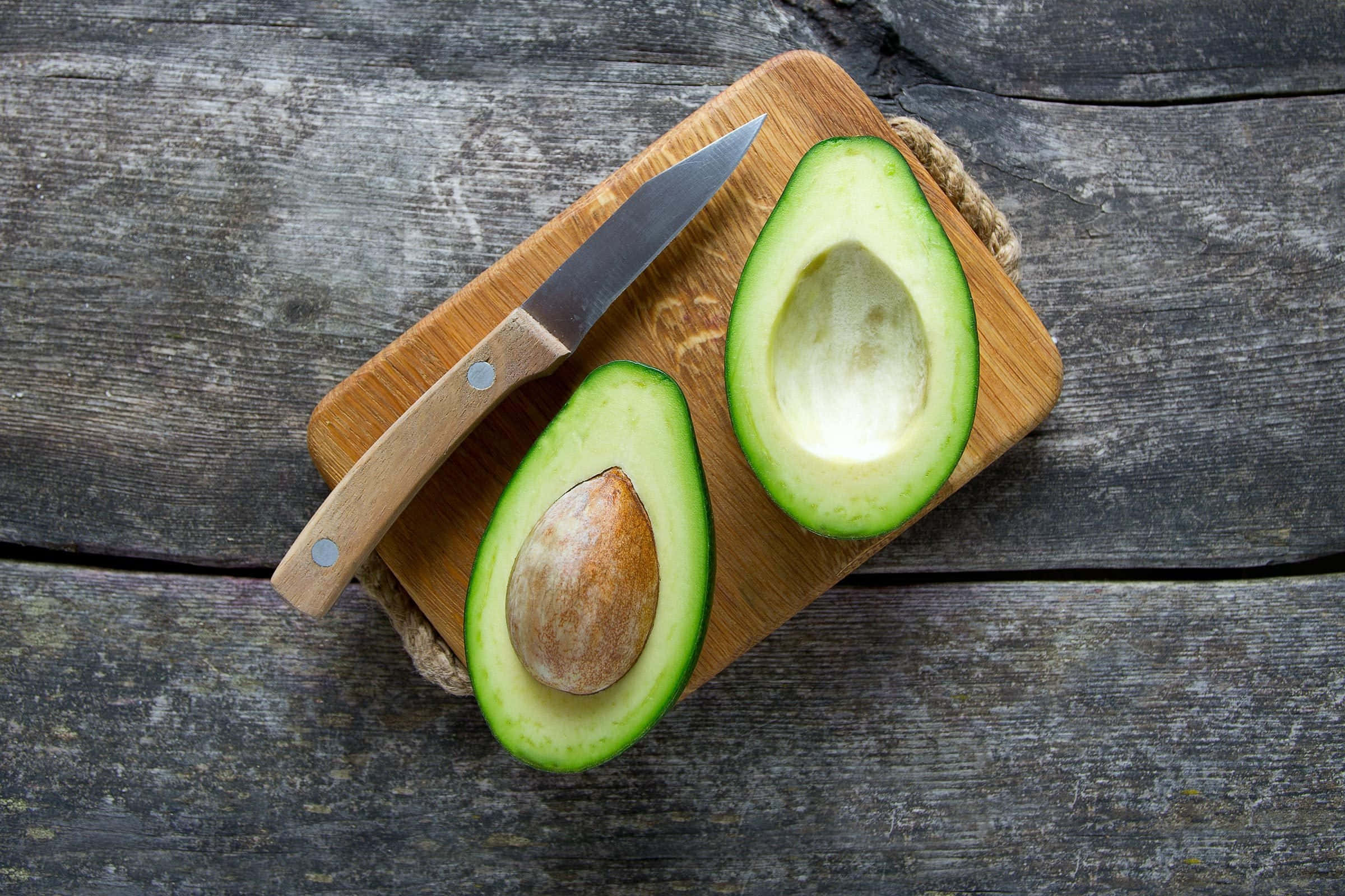 Avocado 桌布 壁紙 — 半個牛油果（有核）放在木砧板上，旁邊有一把刀，置於風化灰色木面上。