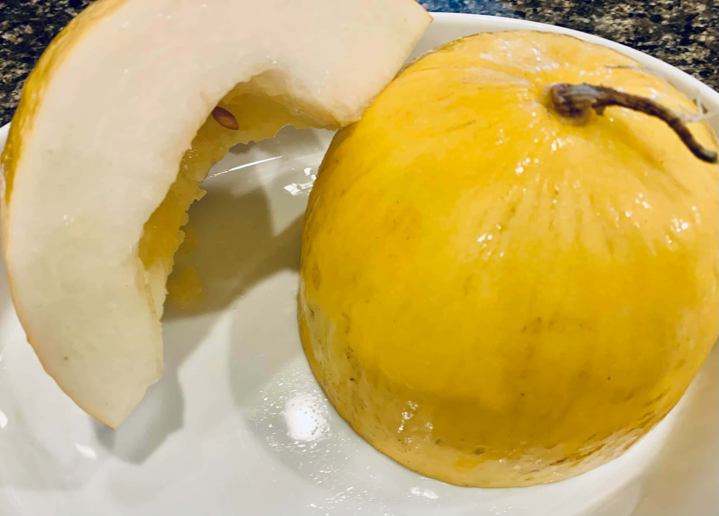 半切黃色Crenshaw蜜瓜 壁紙 — 一個多汁的黃色蜜瓜，切去一片，放在白色碟子上，背景是深色有斑點的枱面。