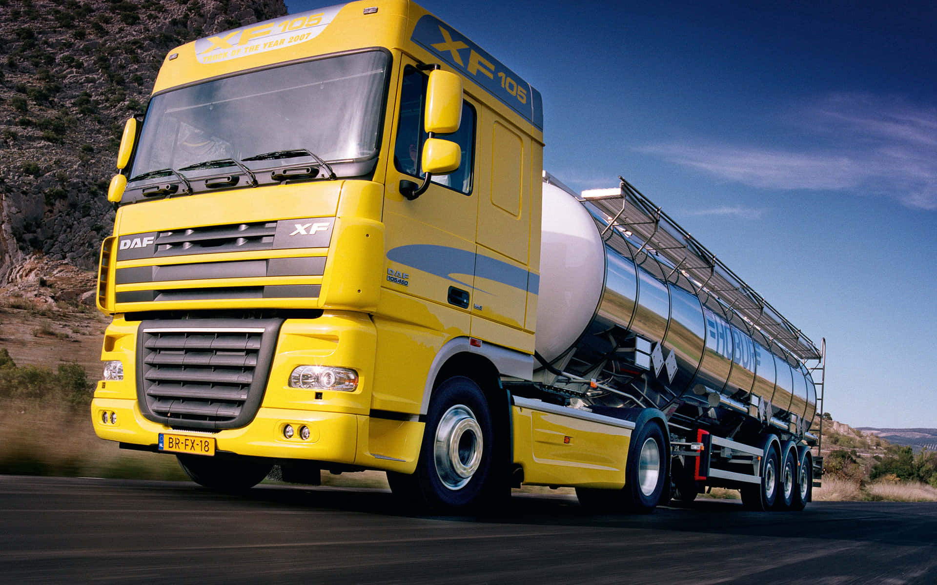 半卡車駛過鄉間彎道 壁紙 — 一輛亮黃色的DAF XF105油罐車，帶有銀色油罐，在晴朗藍天下行駛在乾燥丘陵的道路上。